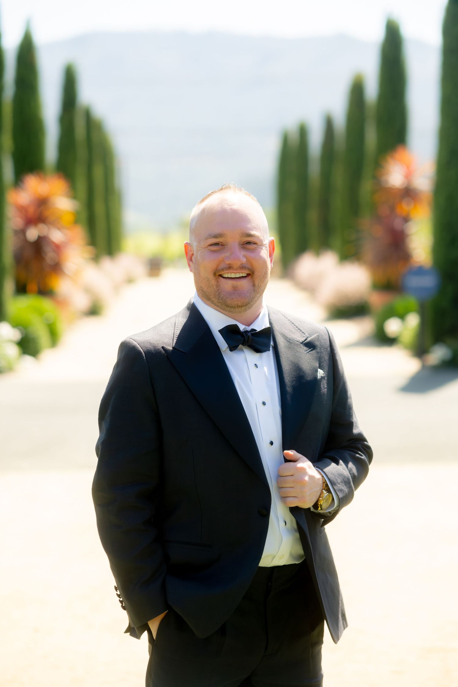 Smiling groom in black tuxedo and bow tie poses outdoors among cypress trees at vineyard wedding.
