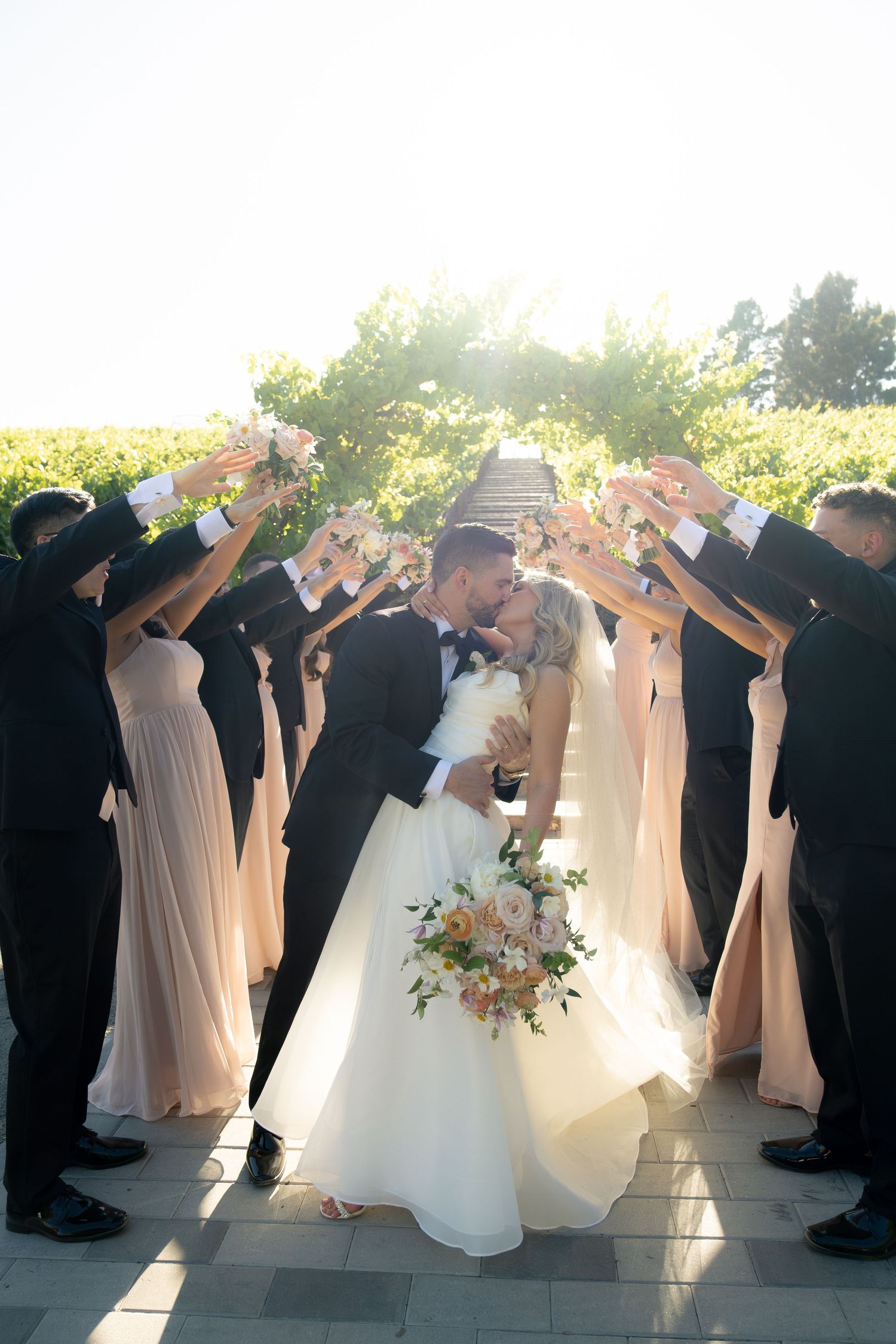 Wedding party raises flowers as bride and groom share a romantic kiss in sun-drenched outdoor ceremony.