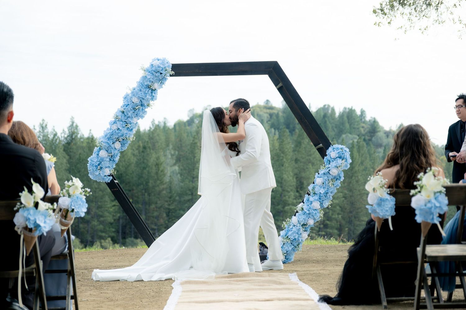 Bride and groom share first kiss under hexagonal arch adorned with blue hydrangeas at outdoor wedding ceremony.