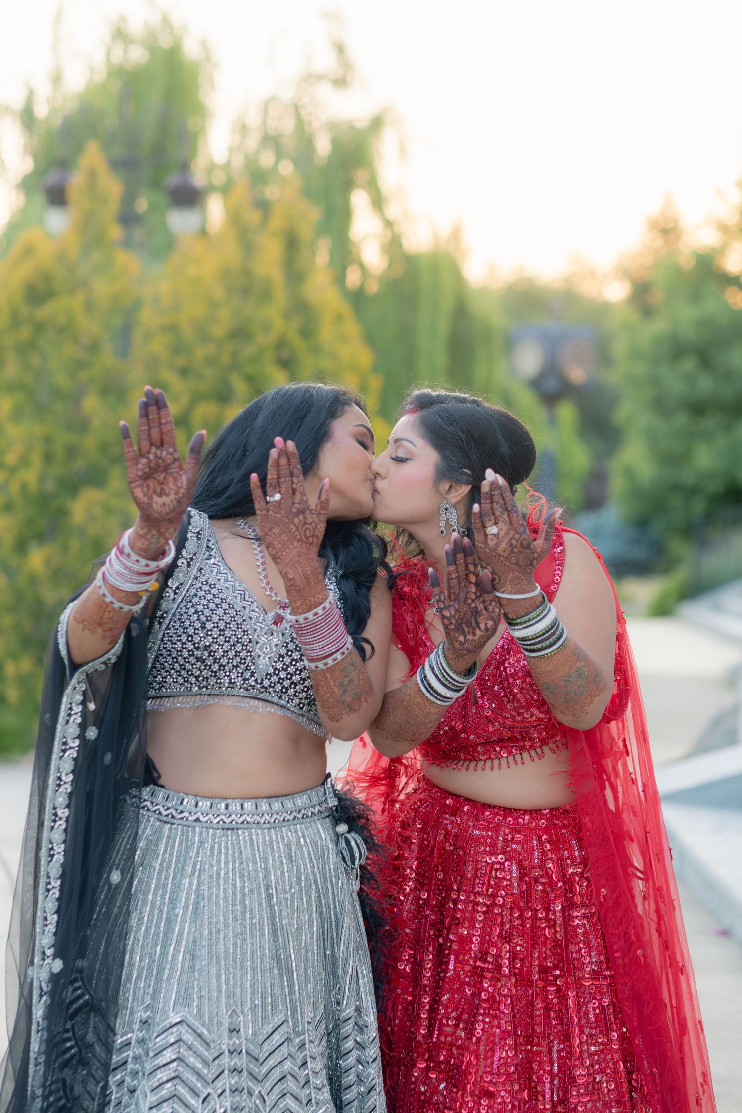 Two Indian women in traditional lehengas kiss cheeks while showing intricate mehndi hands outdoors.