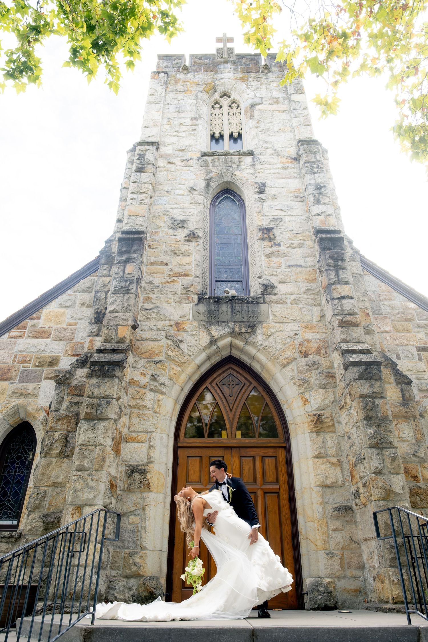 Bride and groom share a romantic dip kiss on the steps of a stone Gothic church.