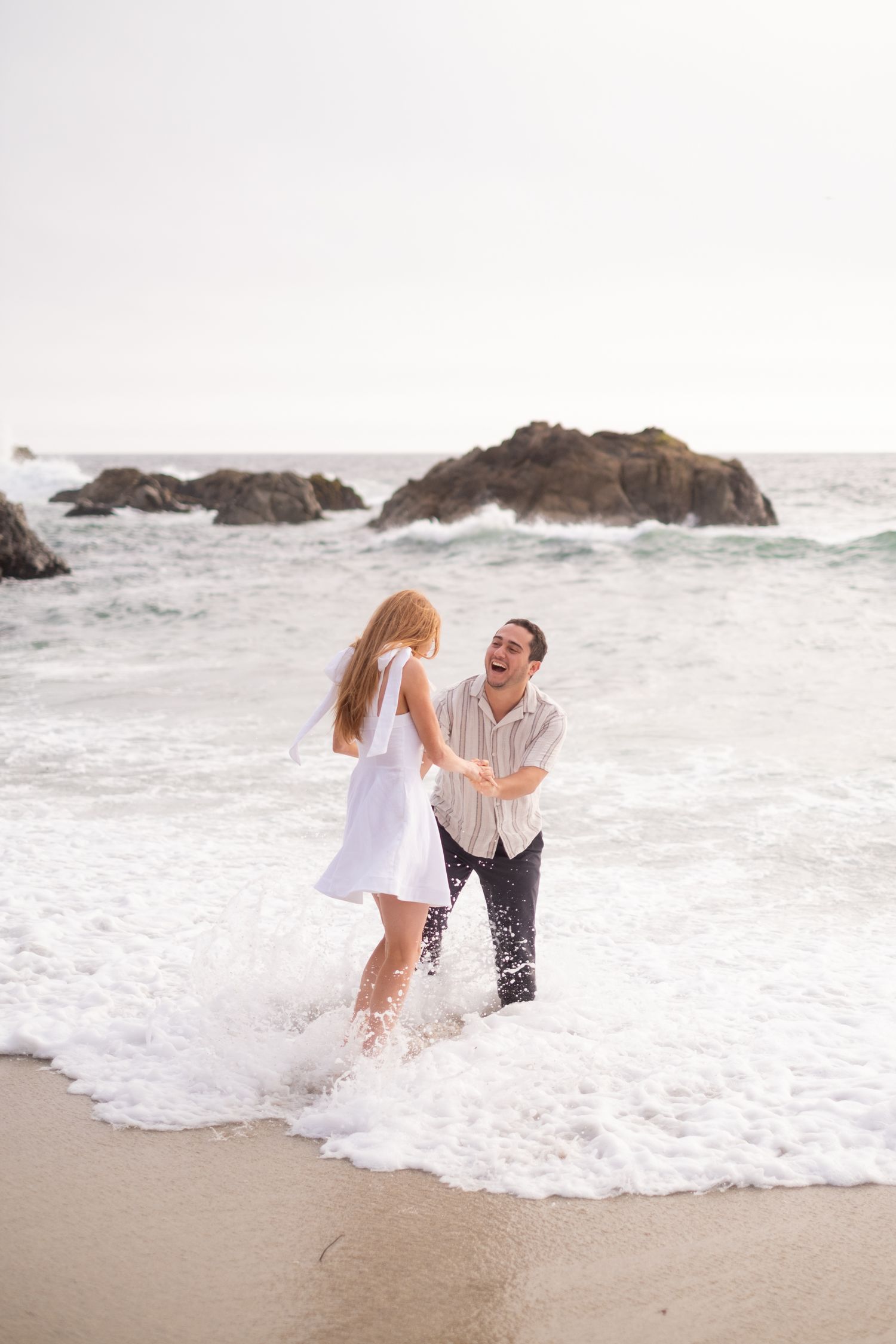 Couple laughing and playing in ocean surf on sandy beach with rocky outcroppings behind them.