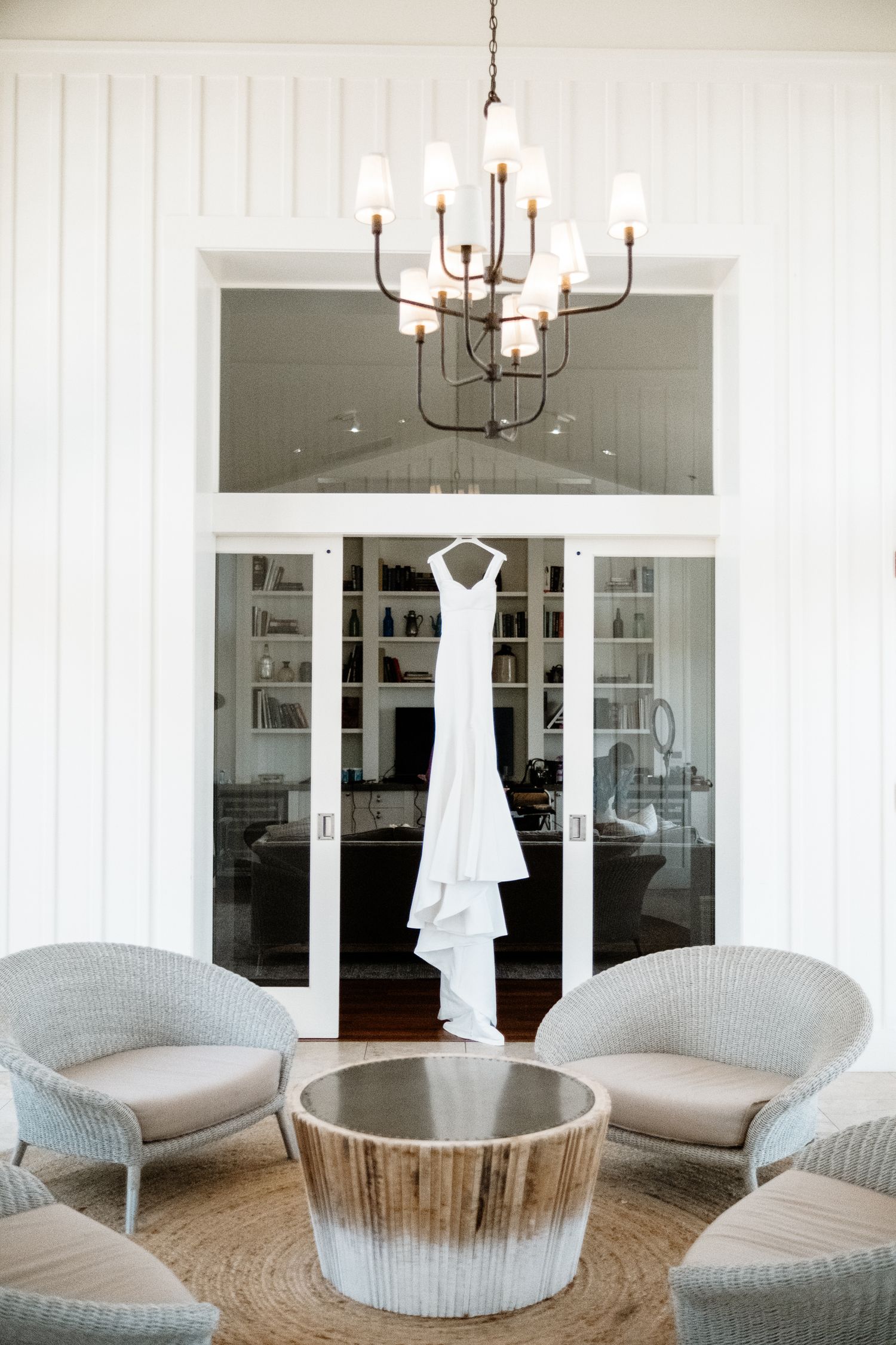 Elegant white wedding dress hanging in doorway beneath chandelier with wicker chairs and round table.
