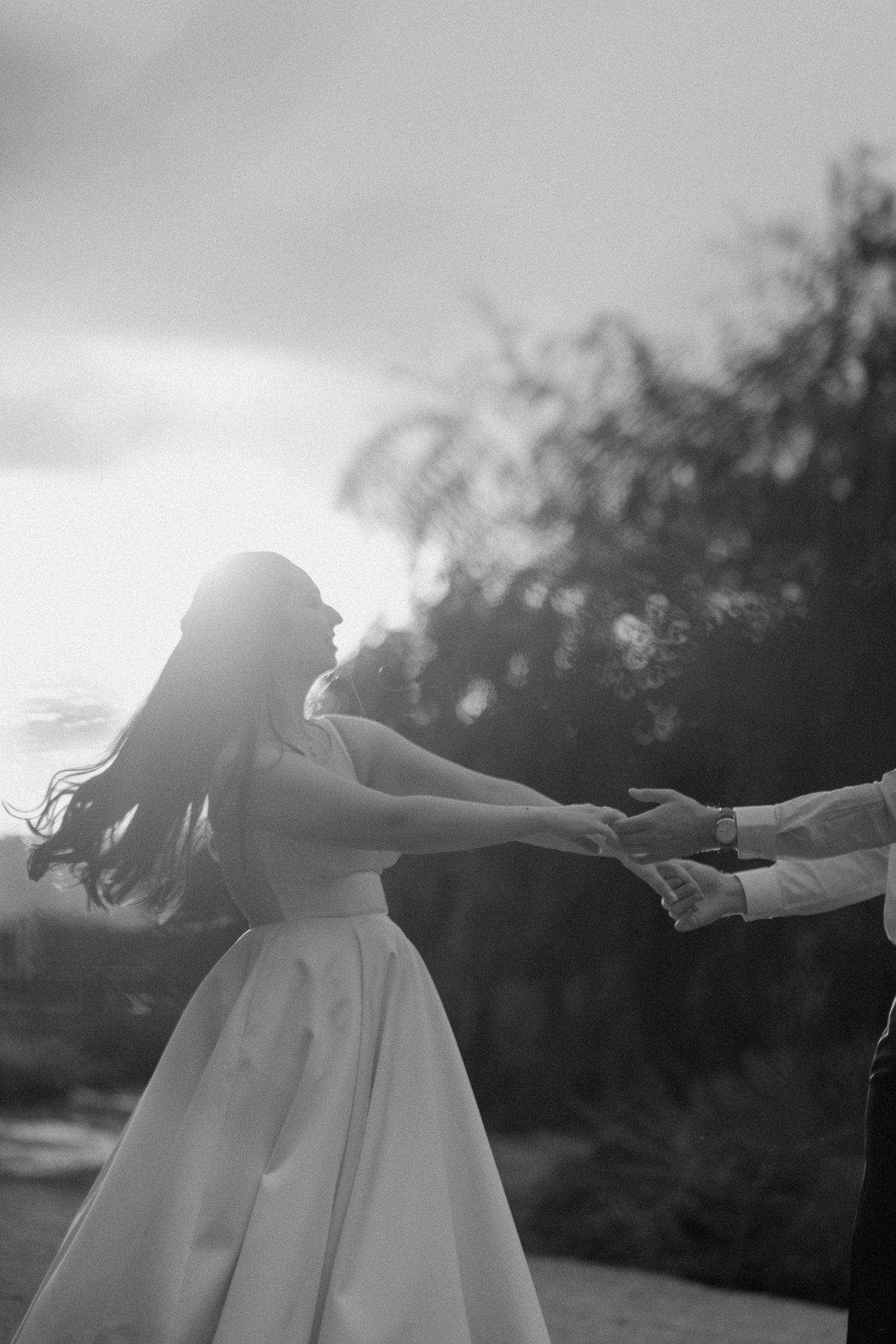 Black and white photo of bride reaching for groom's hands at sunset.