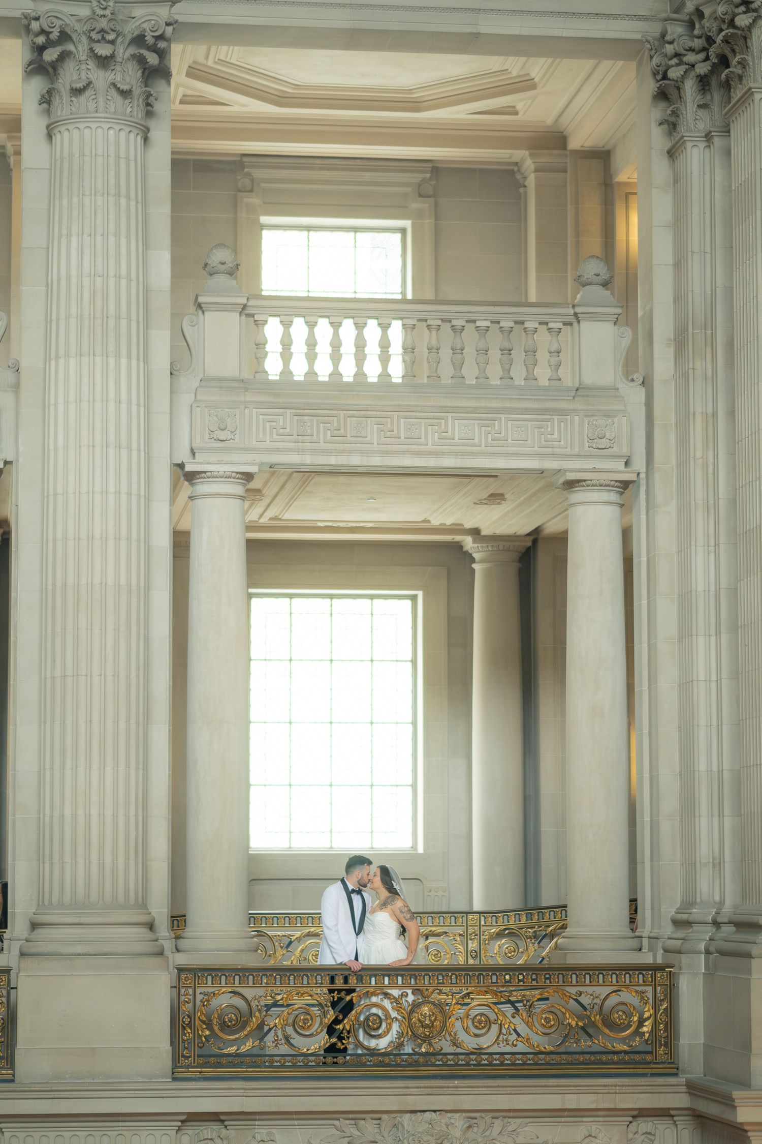 Wedding couple shares romantic kiss on ornate balcony of grand classical architecture venue.