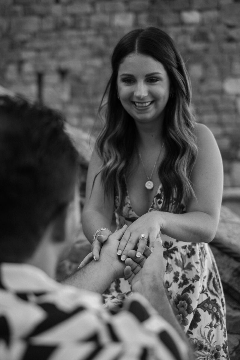 Black and white photo of a smiling woman showing her engagement ring to her partner during a proposal.