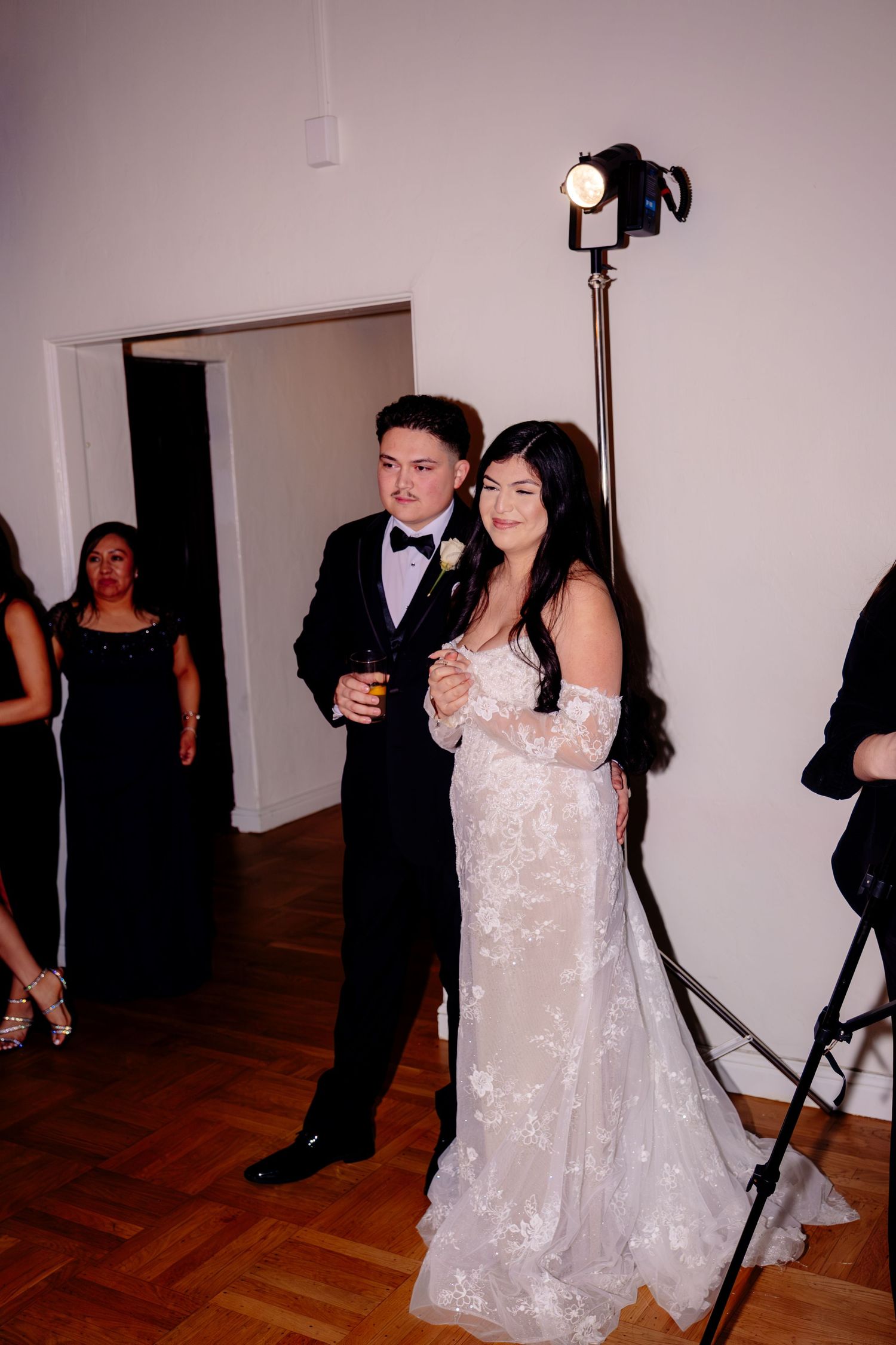 Bride in lace gown and groom in black tuxedo pose together at indoor wedding reception under bright studio light.