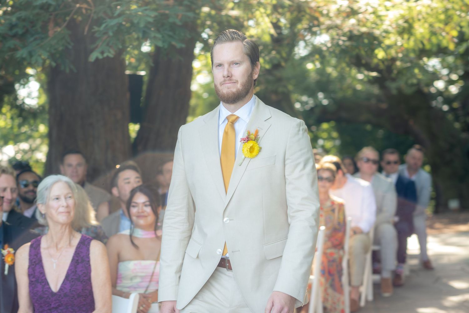 Groom in beige suit with yellow tie and boutonniere walks down outdoor wedding aisle lined with seated guests.