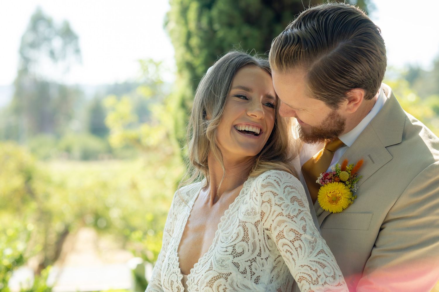 Happy bride in lace dress laughing with groom in tan suit outdoors at sunny vineyard wedding ceremony.