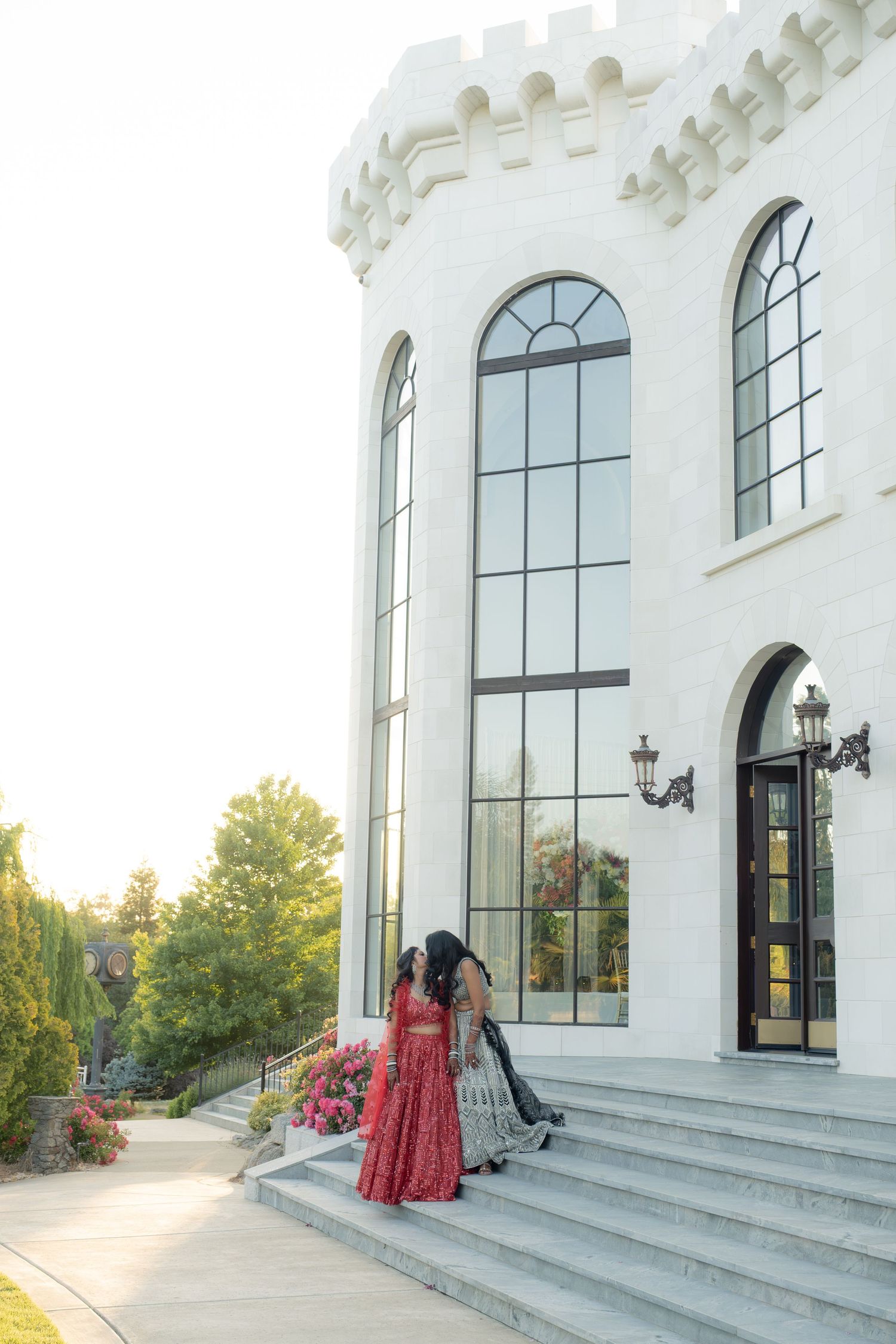 Two women in colorful Indian attire on steps of white castle-like wedding venue at golden hour.