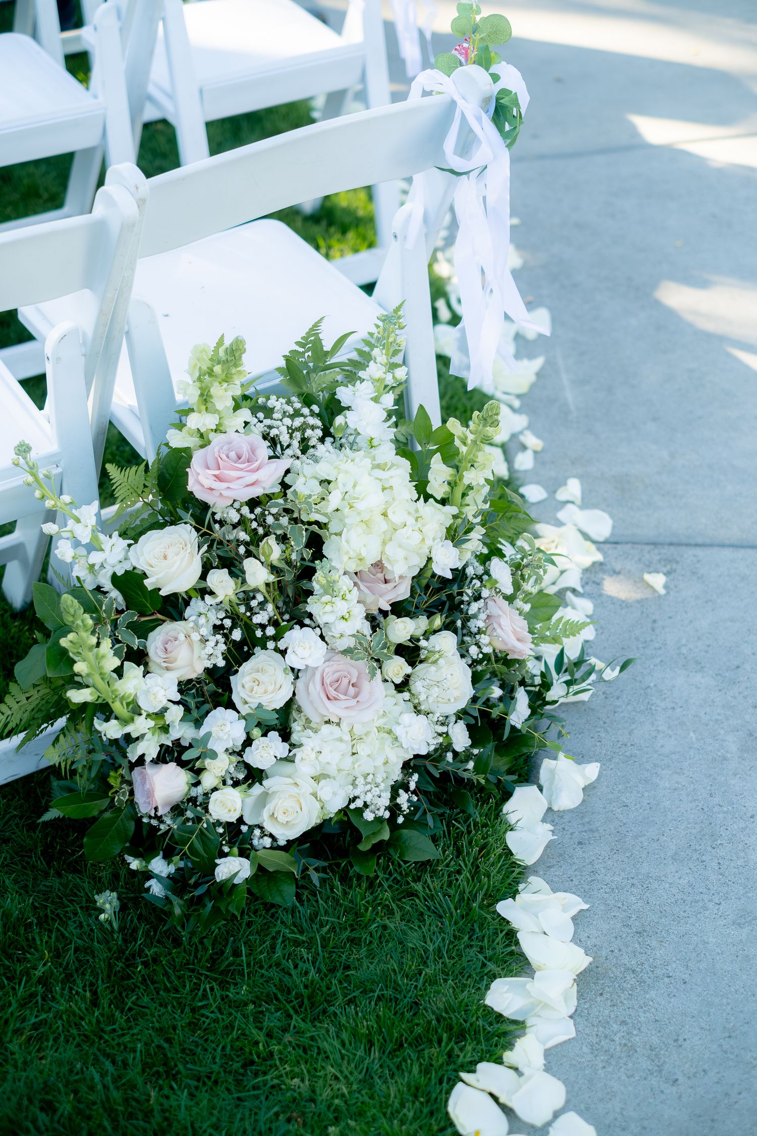 Elegant wedding aisle with white chairs adorned with ribbon bows and lush floral arrangements of pink roses and white blooms on green grass.