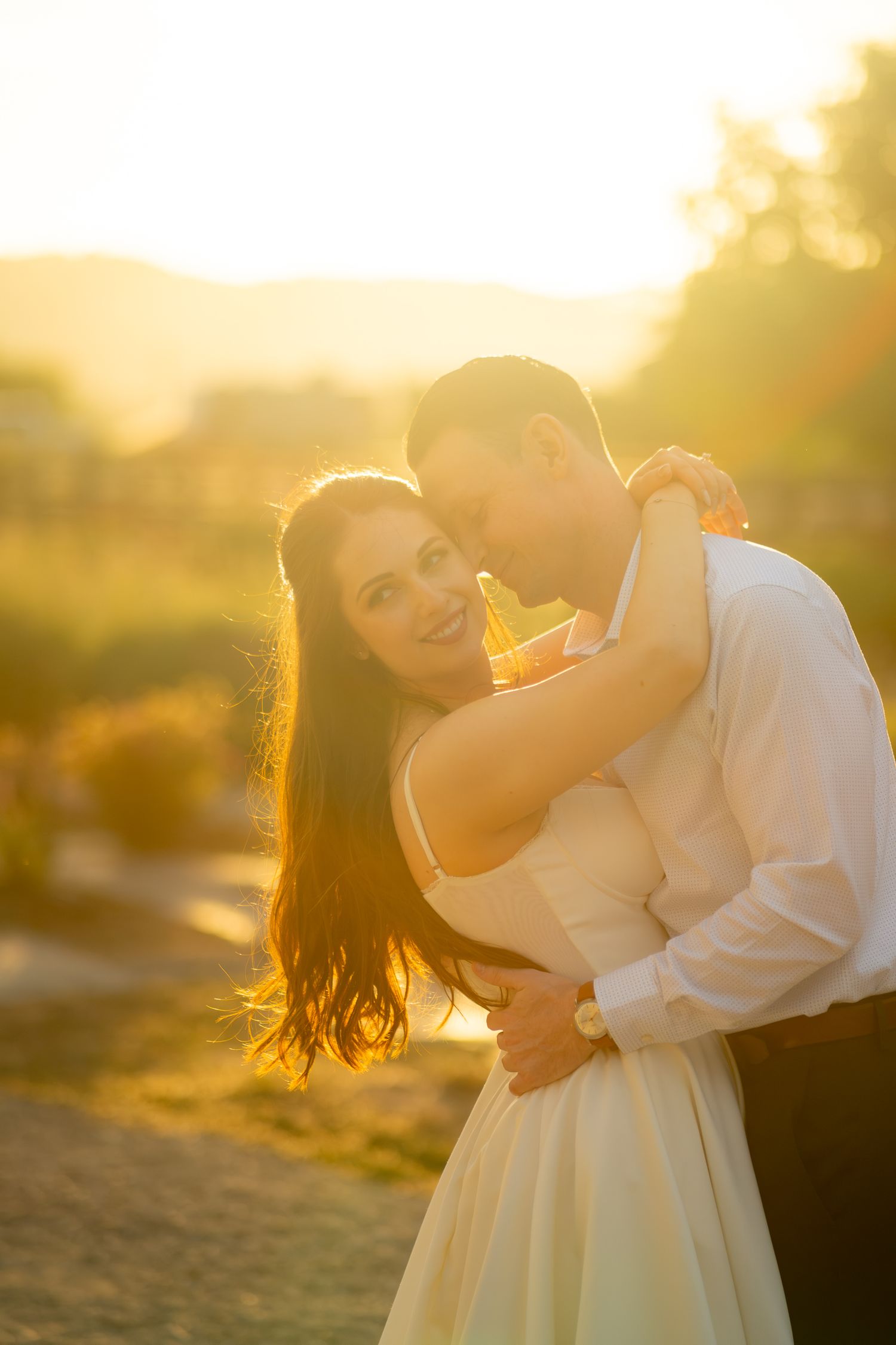 Couple embracing at golden hour sunset, bride in white dress smiling as groom kisses her cheek outdoors.
