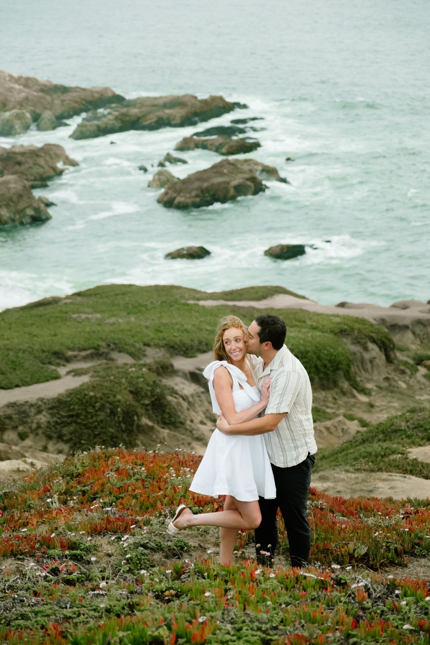 Couple embracing on a coastal hillside with wildflowers and rocky ocean shoreline in the background.