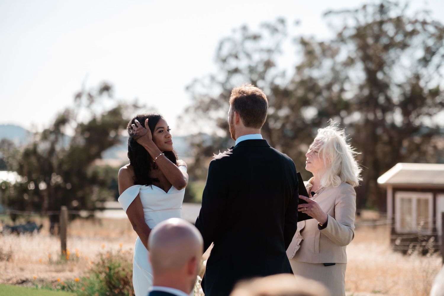 Bride and groom exchanging vows outdoors with officiant during sunny wedding ceremony in natural setting.