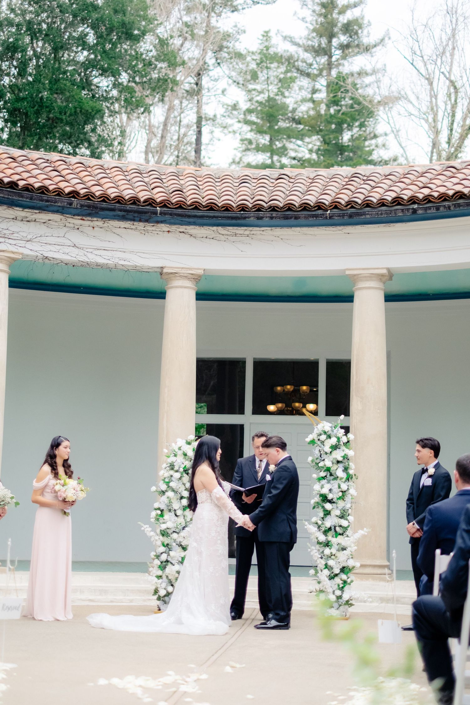 Bride and groom exchange vows at a floral arch altar before a classical columned pavilion with terracotta roof.