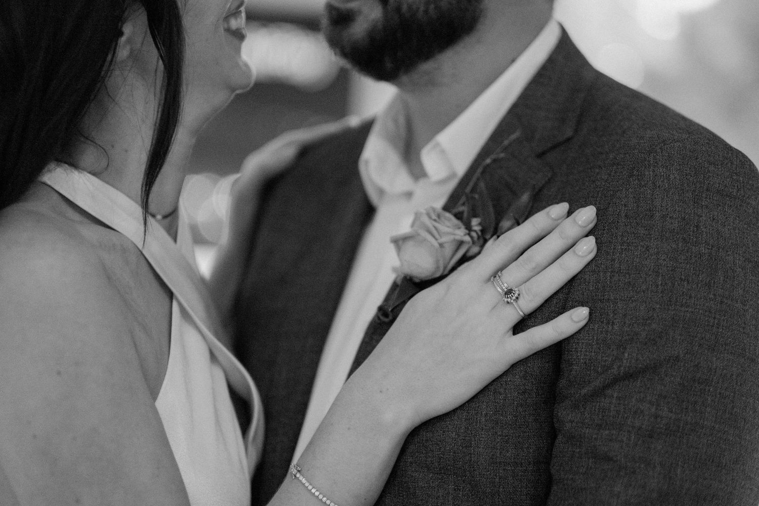 Black and white close-up of bride and groom embracing, showcasing diamond engagement ring and floral boutonniere.