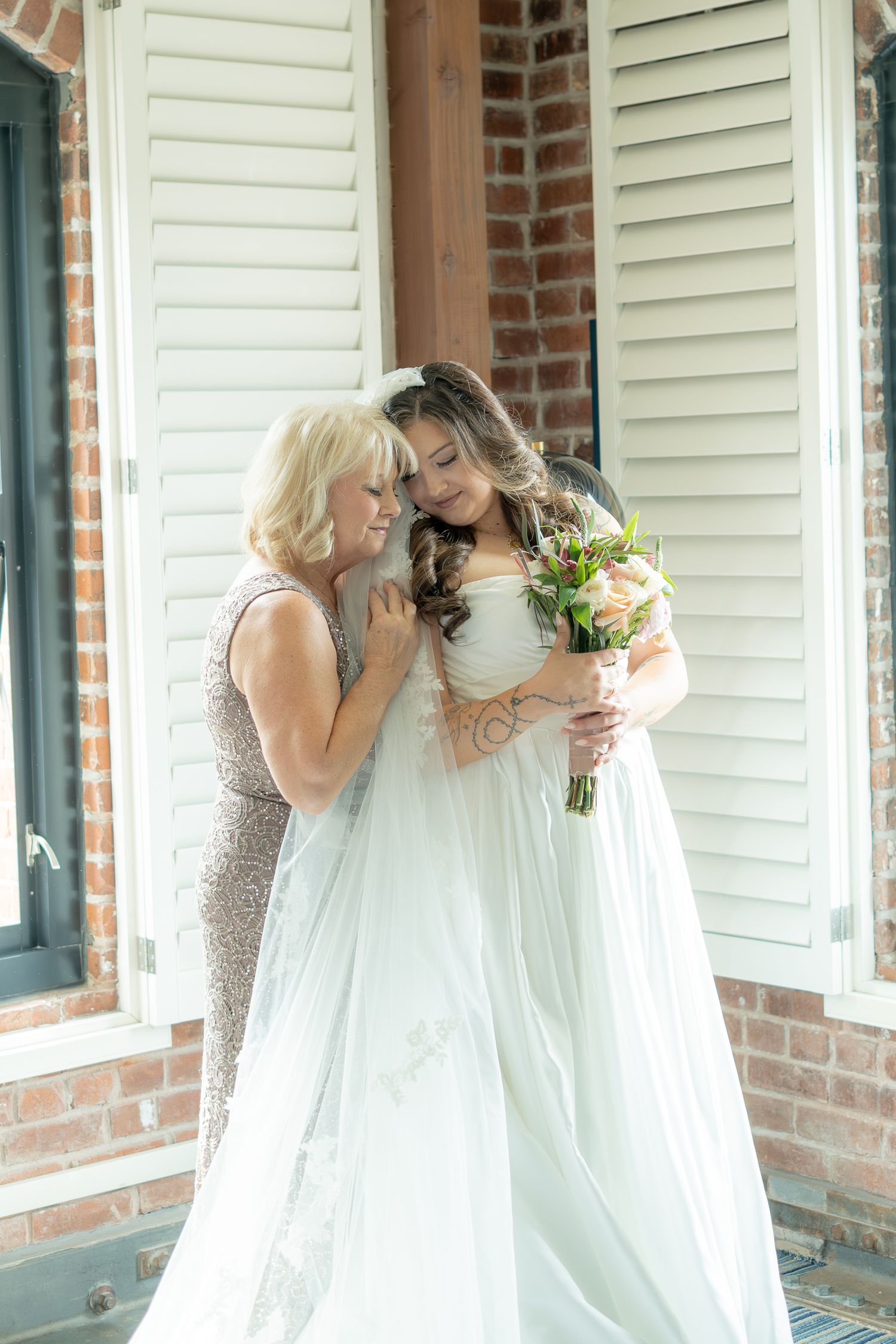 Bride in white gown holding bouquet shares tender moment with older woman in sequin dress outside brick building.