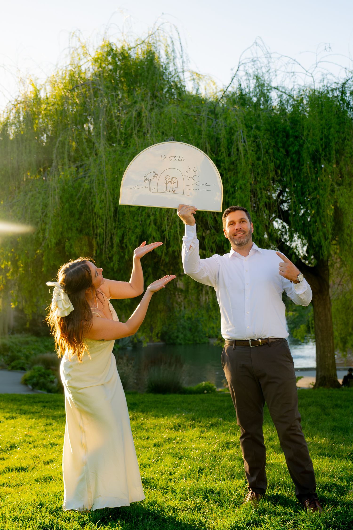 Bride and groom holding a decorative wedding sign outdoors near a willow tree on a sunny day.