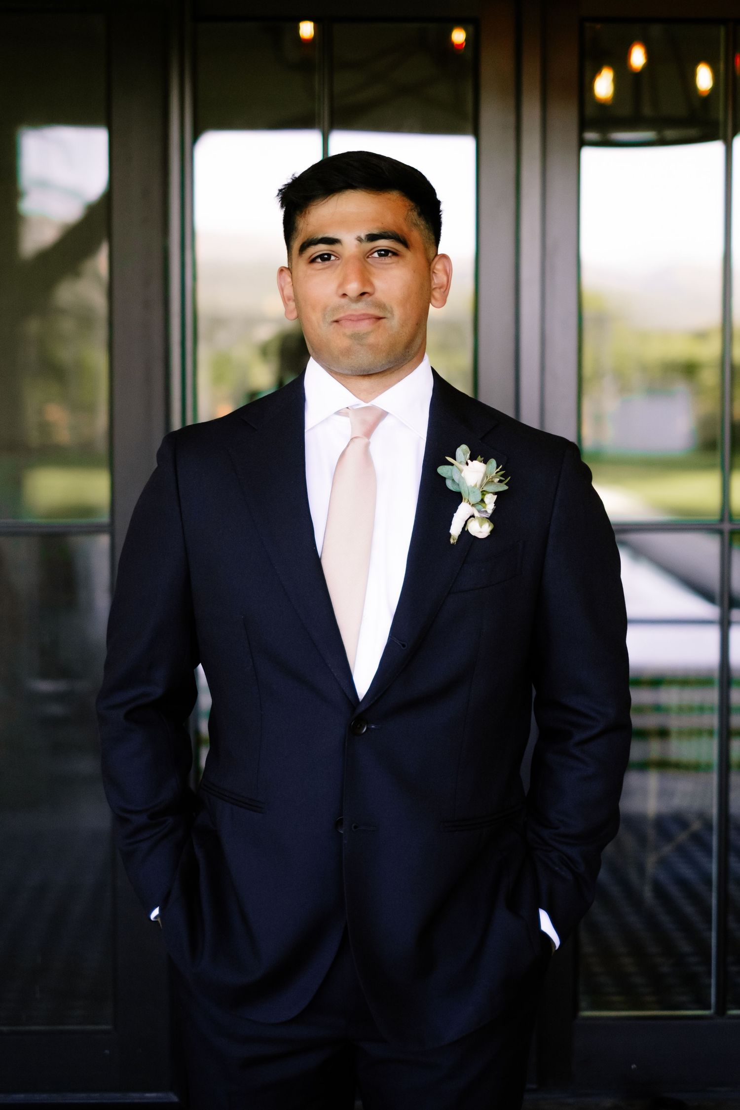 Groom in a navy suit with pink tie and boutonniere standing confidently before glass doors.