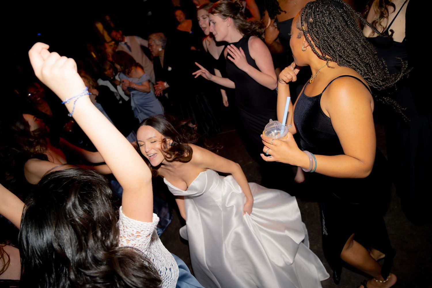 Bride in white gown dancing energetically with guests on crowded dark wedding reception dance floor.