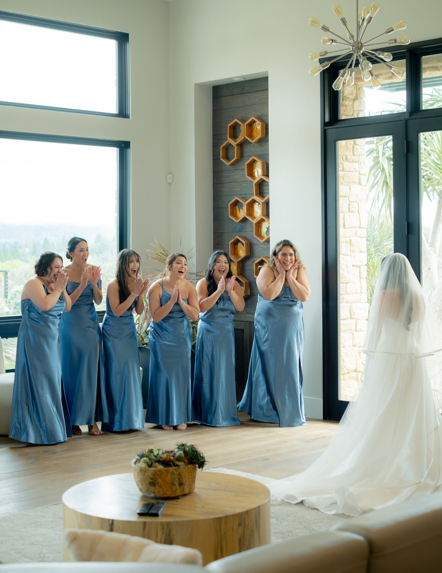 Bridesmaids in blue satin gowns react with joy during a first look reveal in an elegant modern venue.