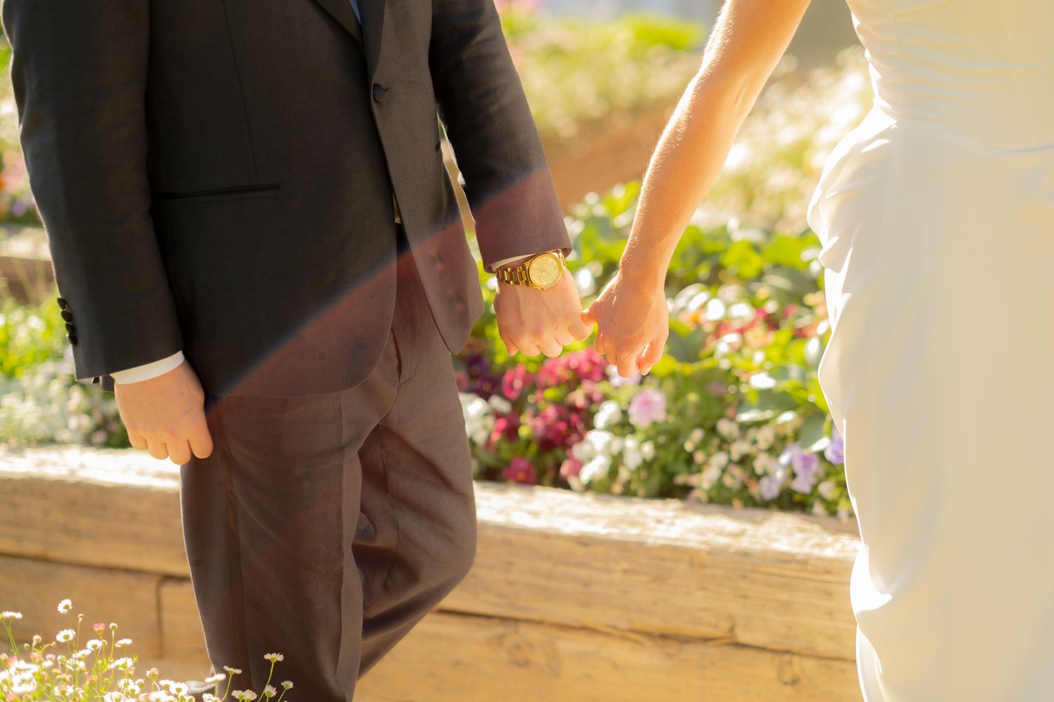 Close-up of bride and groom holding hands with colorful flower garden in background during outdoor wedding celebration.