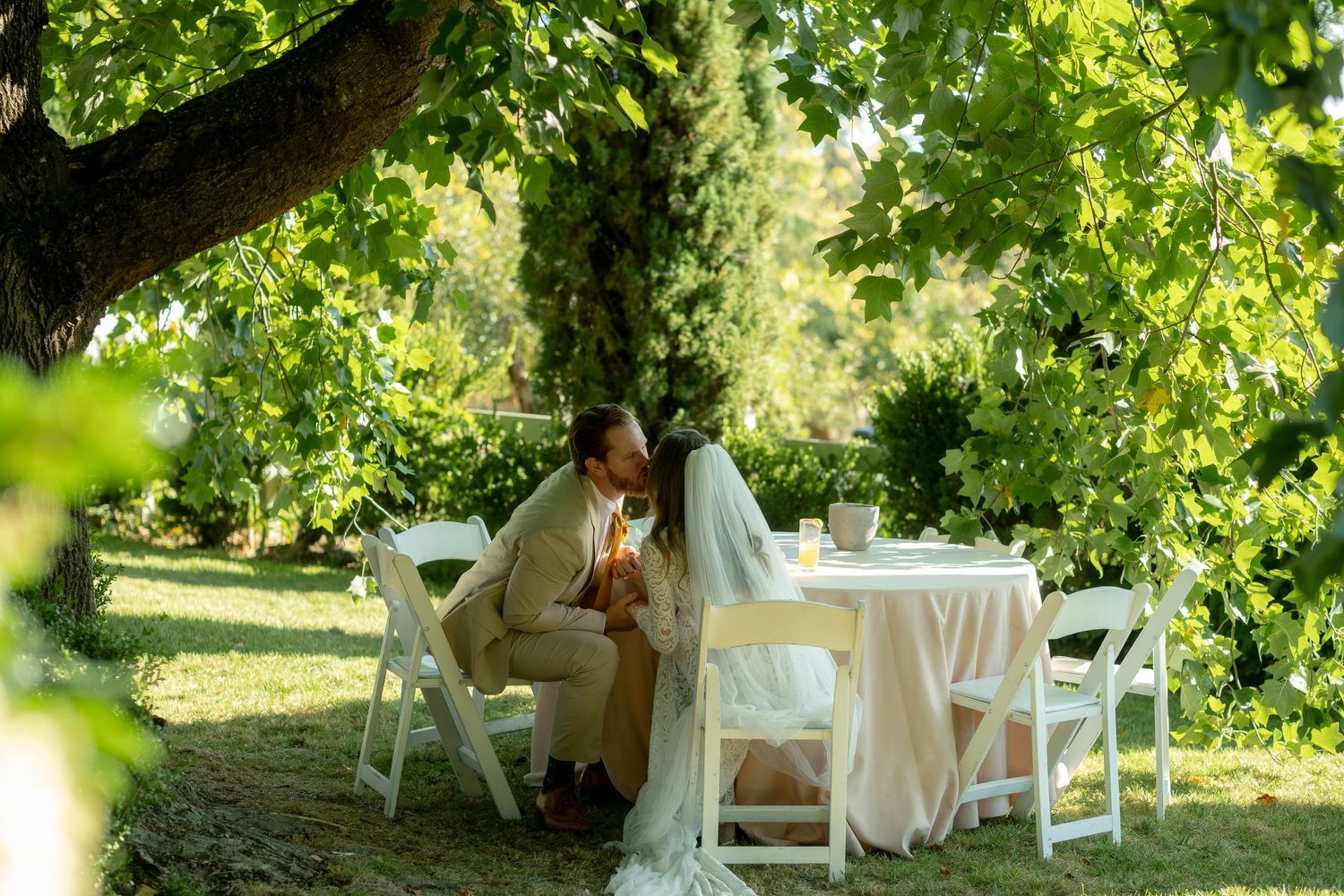 Bride and groom share intimate moment seated at table beneath lush green trees at outdoor wedding reception.