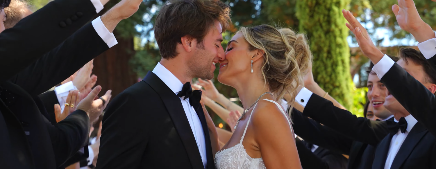Bride and groom share a passionate kiss surrounded by cheering wedding guests in formal attire.