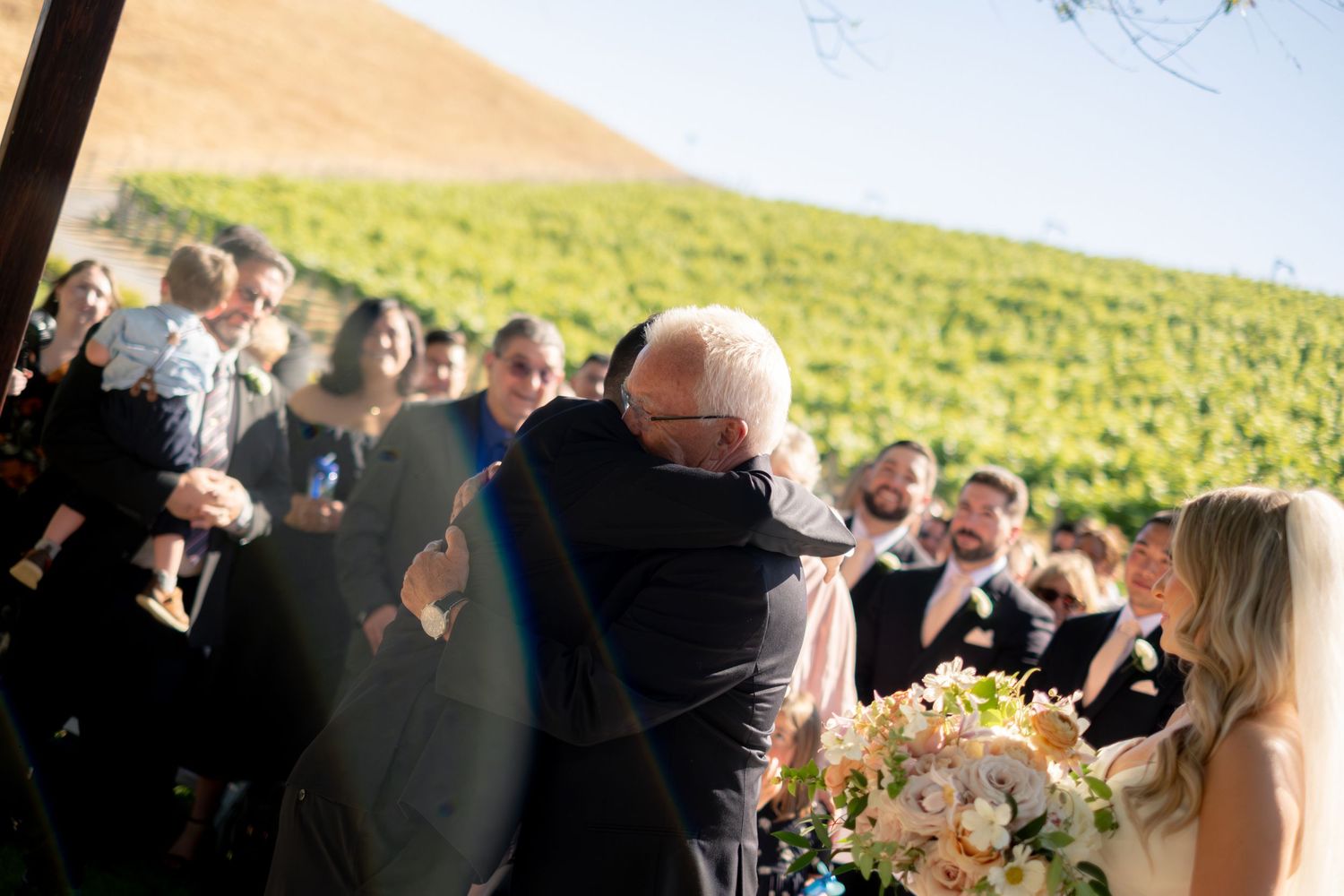 Emotional father-of-the-bride embrace during outdoor vineyard wedding ceremony with guests watching.