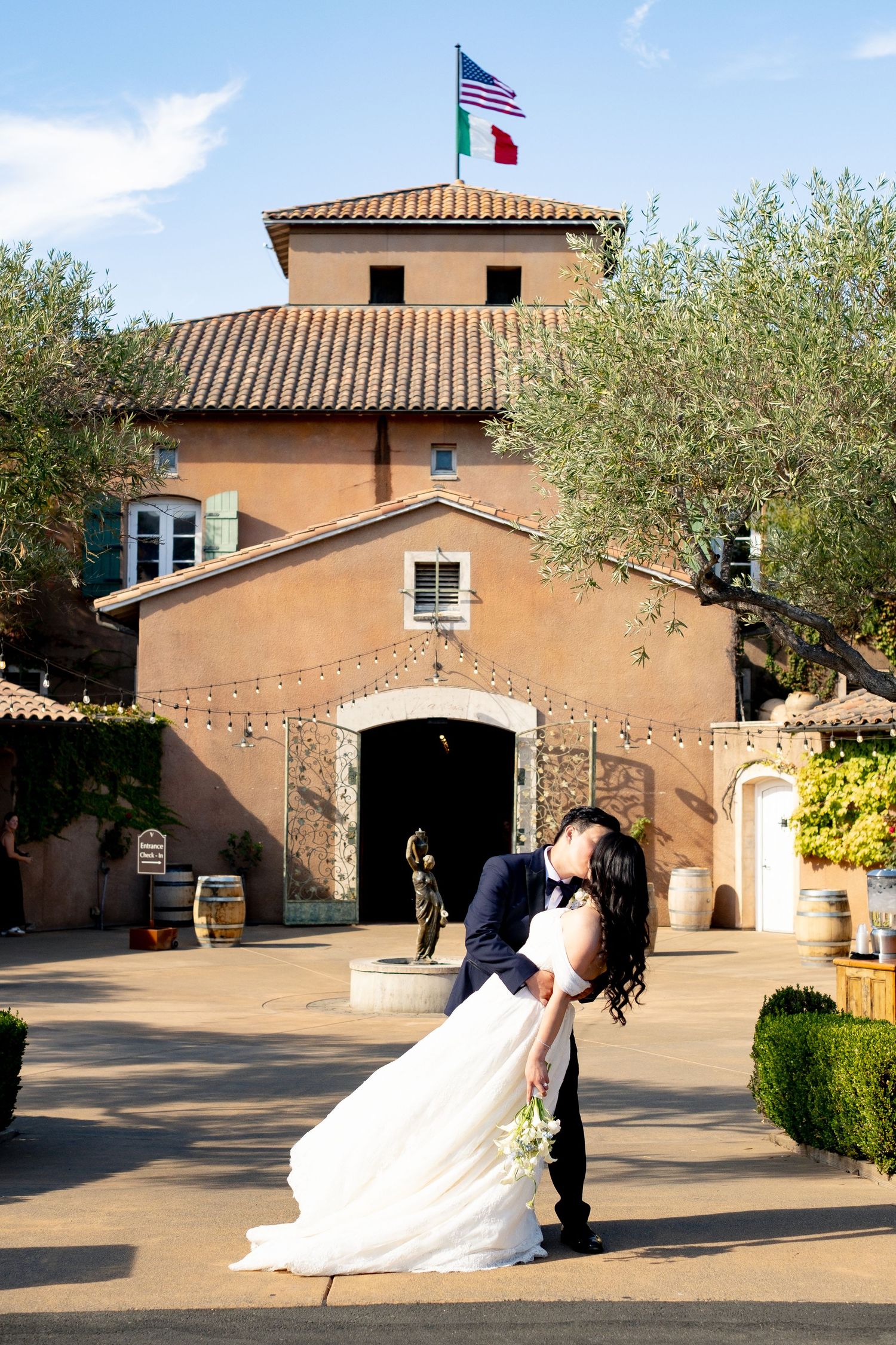 Groom dips bride in a romantic kiss outside an Italian-style winery with flags flying above.