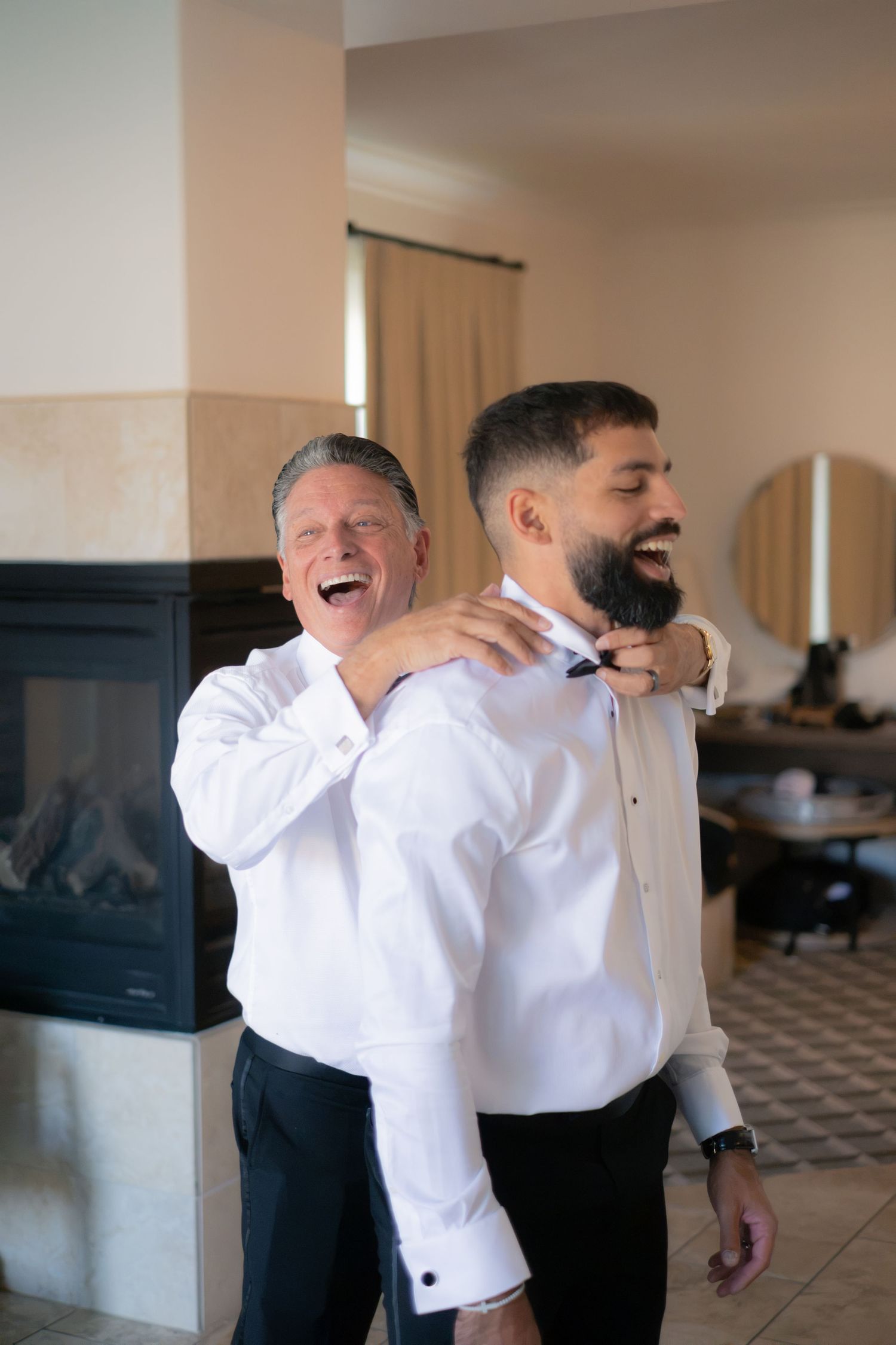 Father and son share a joyful laugh while adjusting bow tie during wedding day preparations indoors.
