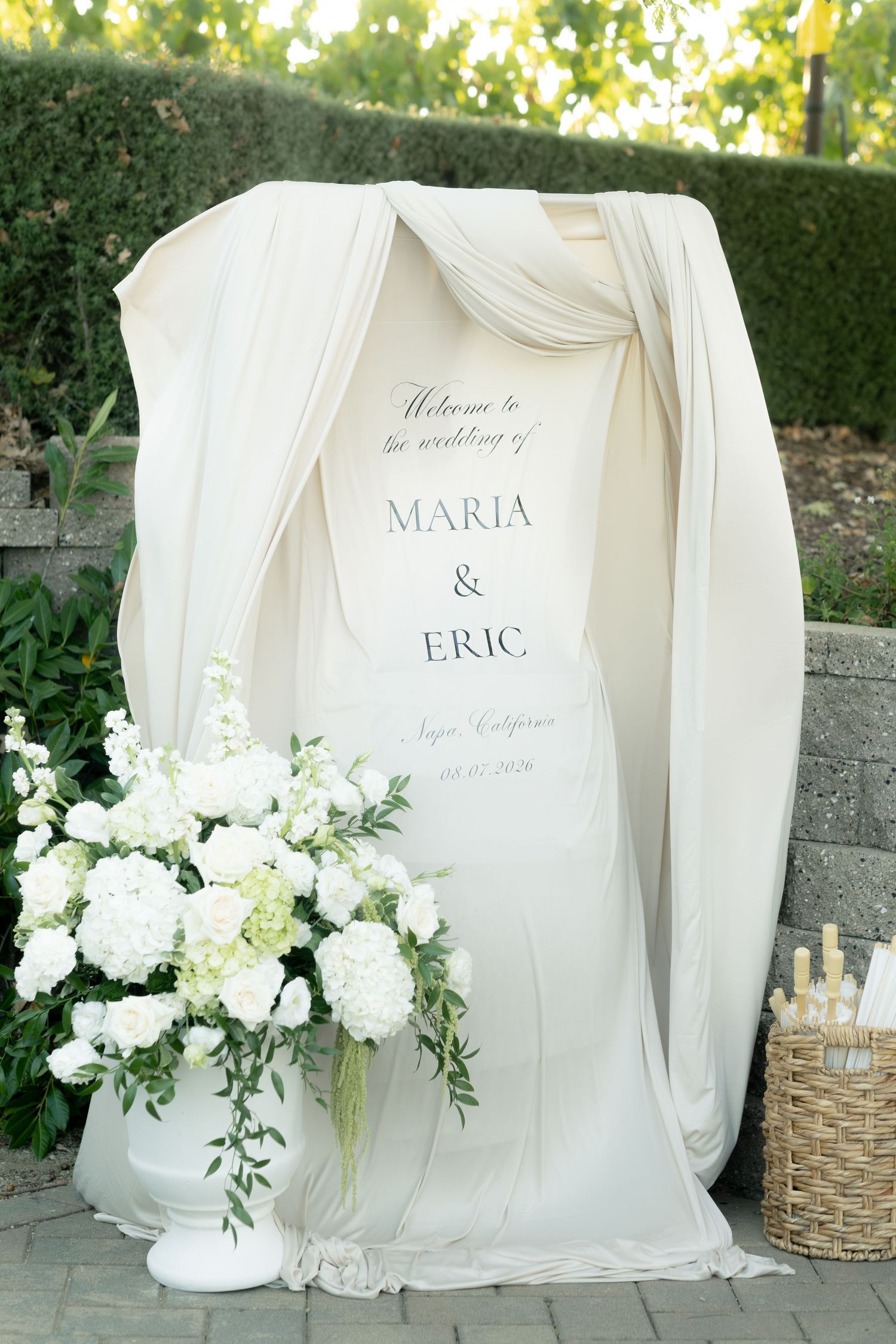 Elegant outdoor wedding welcome sign for Maria and Eric draped in ivory fabric with white floral arrangement.