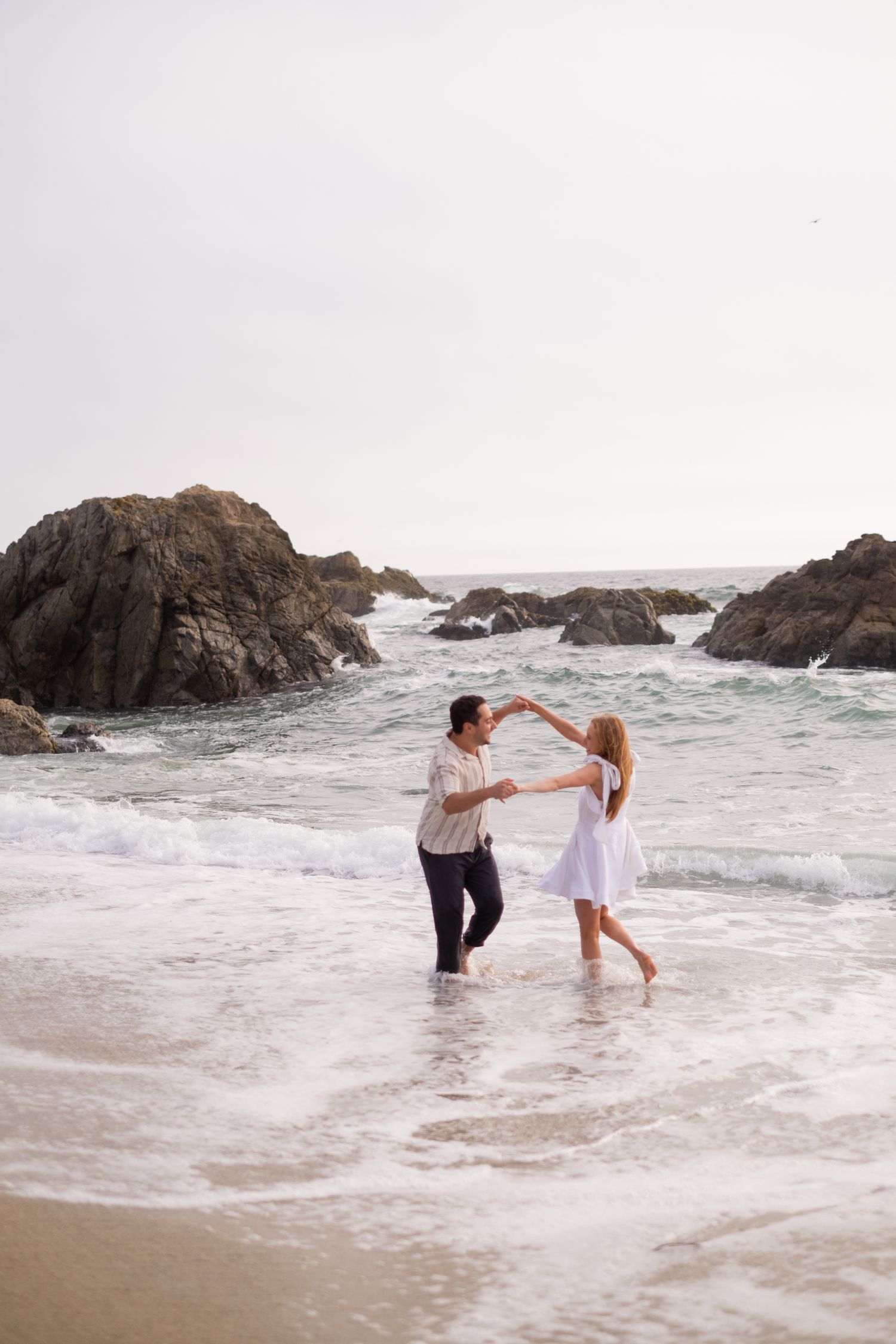 Couple dancing together in shallow ocean waves with large rocks and sea in background.