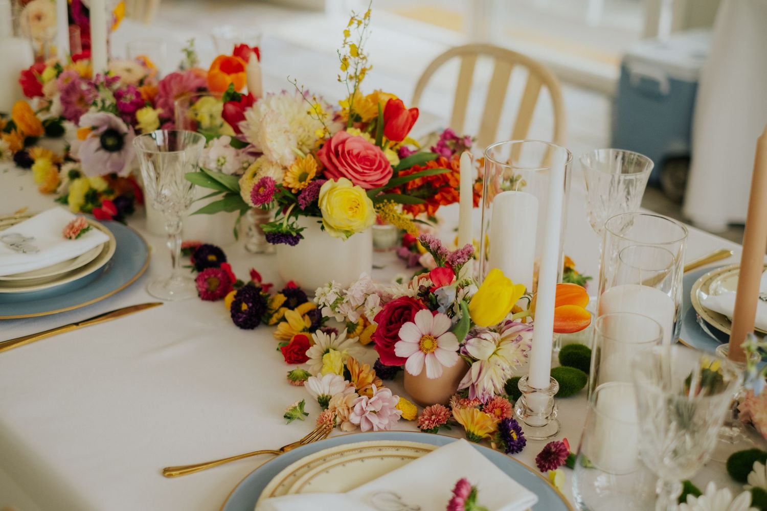 Colorful wedding table centerpiece with vibrant flowers including red, yellow, and pink blooms on a white linen tablecloth.