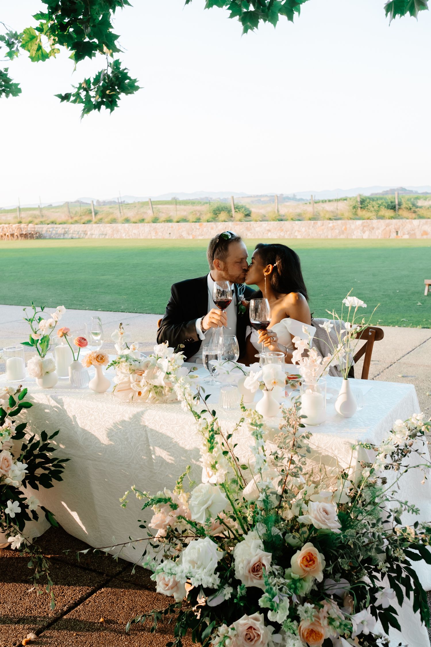 Bride and groom share a kiss at sweetheart table adorned with white and peach floral arrangements at outdoor reception.