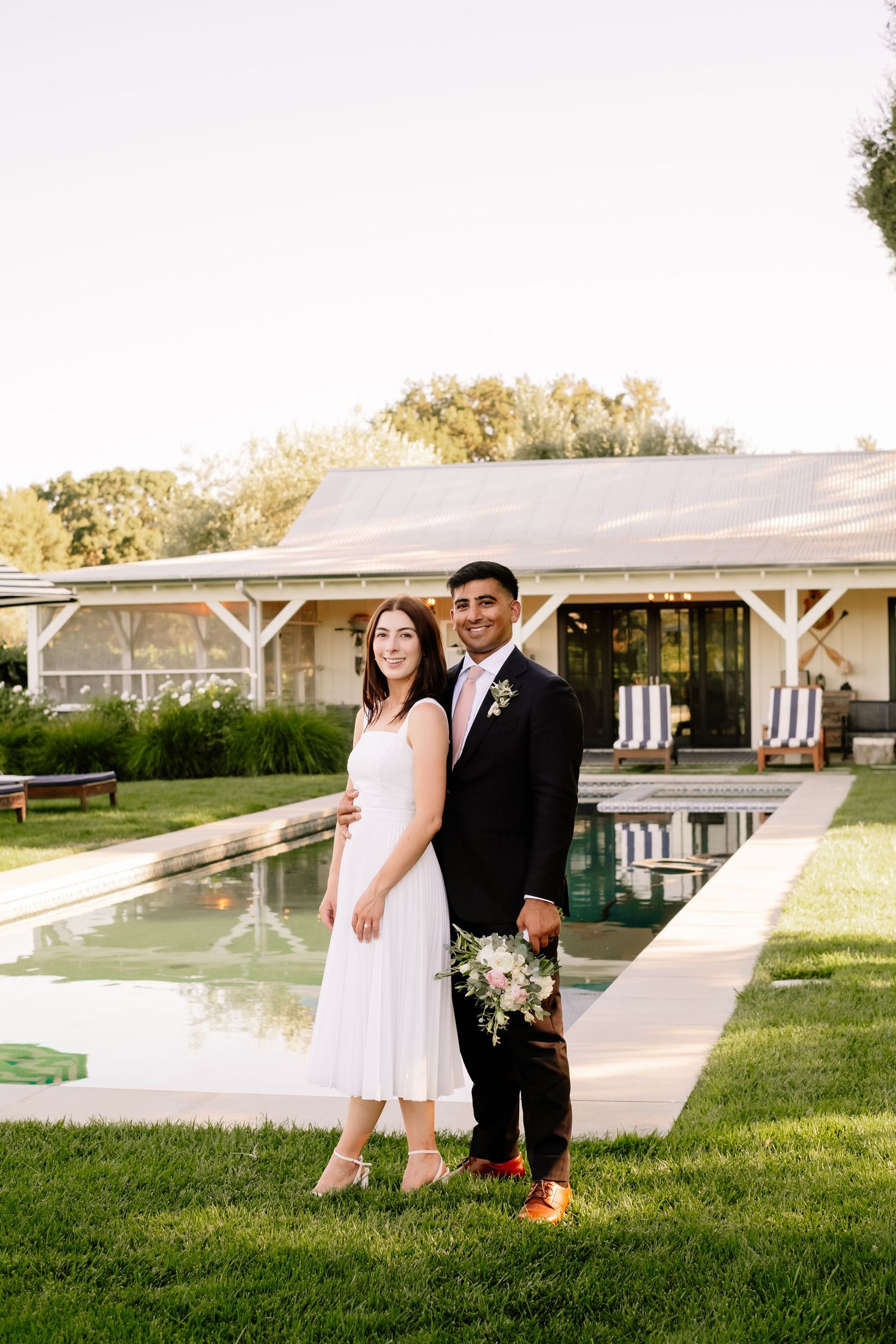 Bride and groom posing together on green lawn beside a pool at their wedding venue.