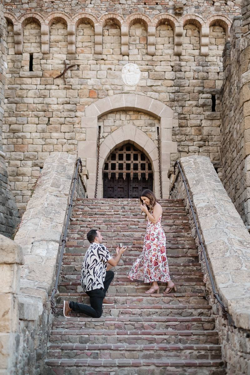 Man proposes on one knee to woman in floral dress on stone castle stairs.