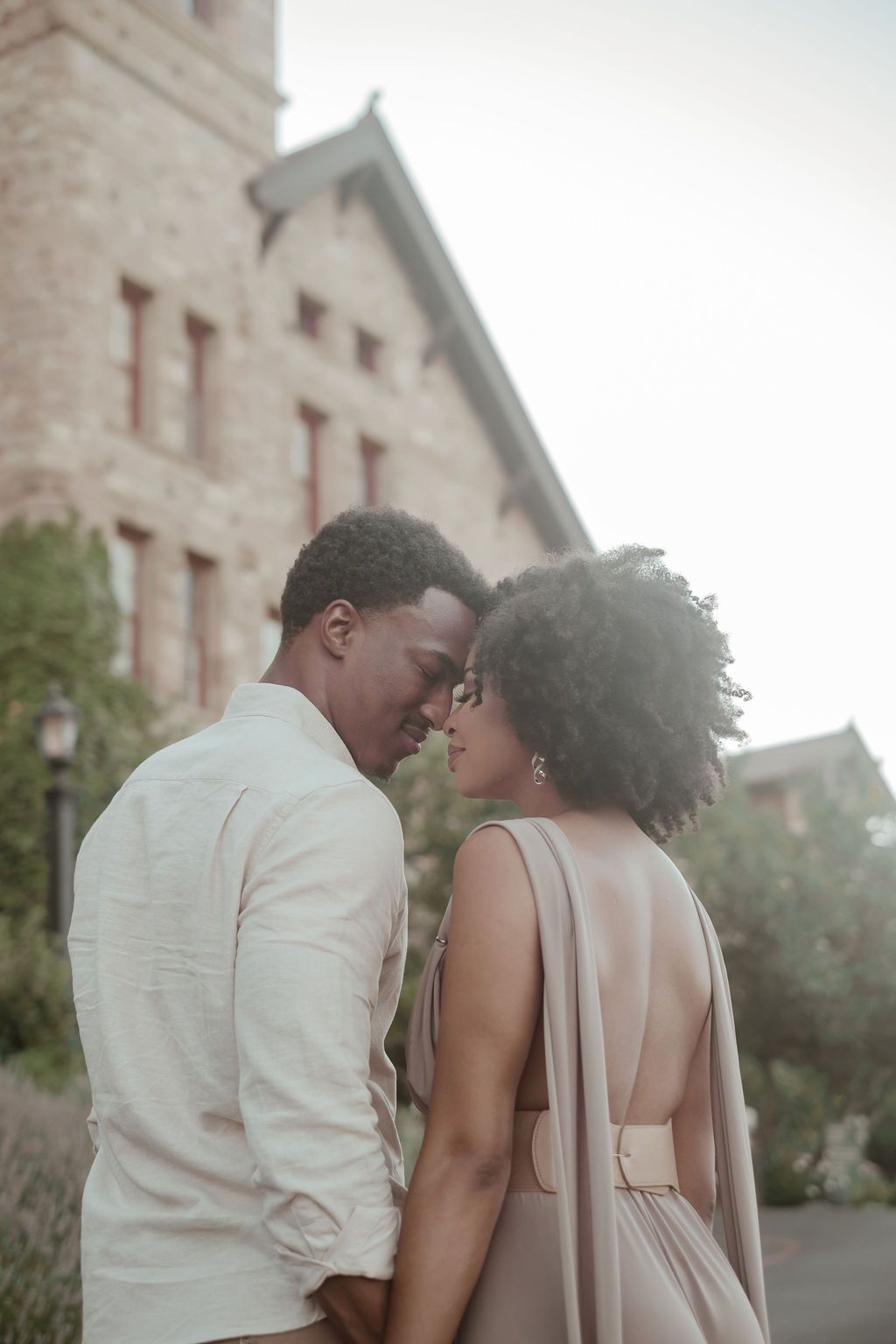 An elegant Black couple leans foreheads together in front of a historic stone building at dusk.