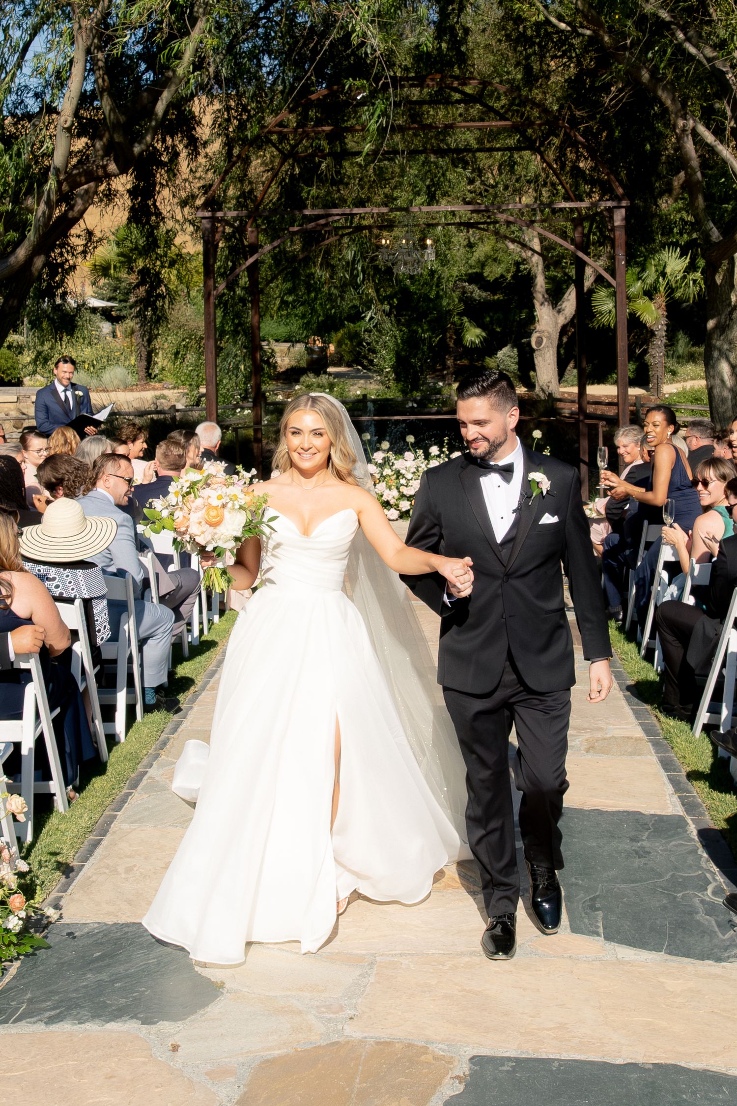 Bride and groom walk down outdoor wedding aisle, smiling joyfully, surrounded by seated guests and lush greenery.