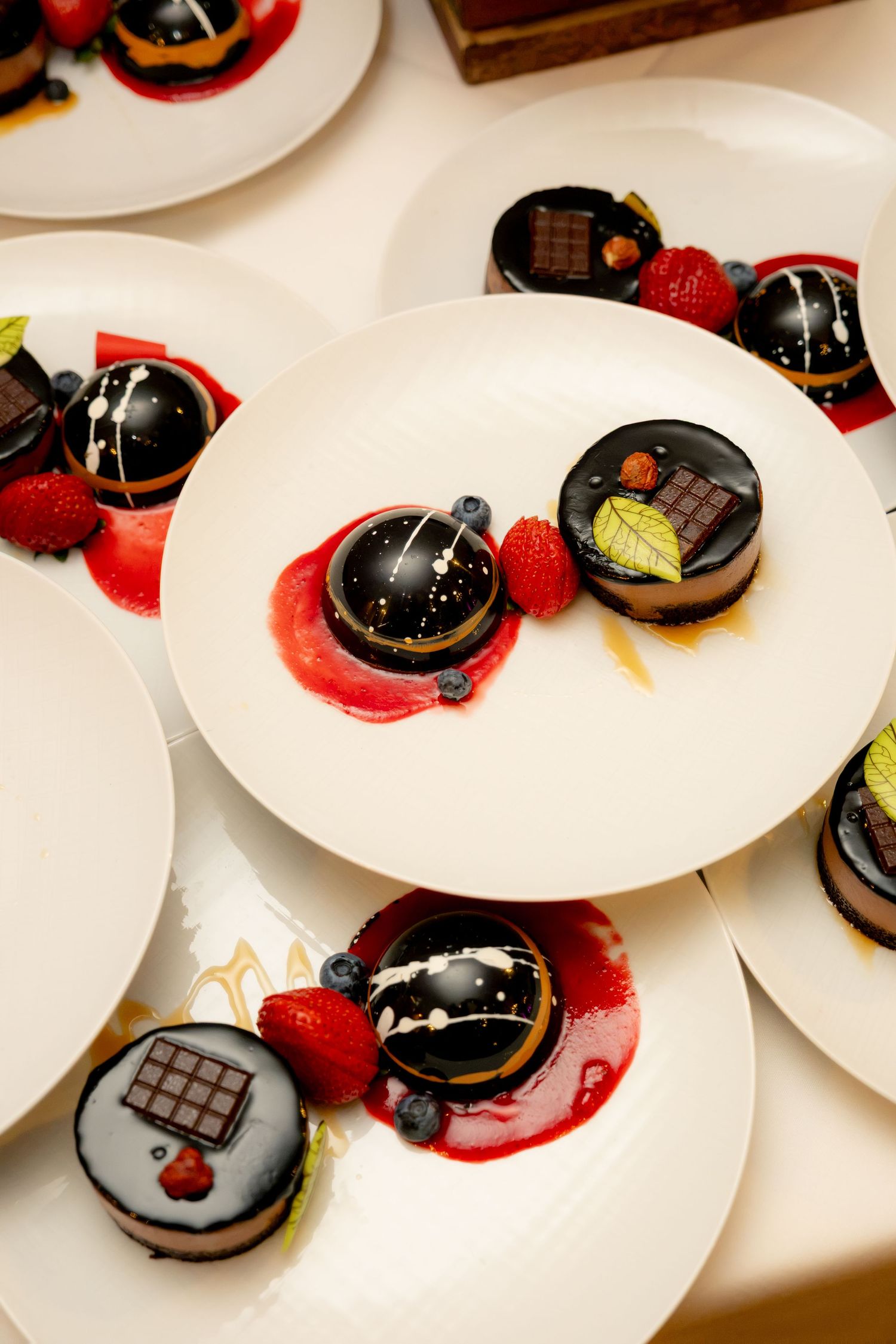 Elegant chocolate mirror glaze desserts garnished with fresh berries and mint served on white plates.