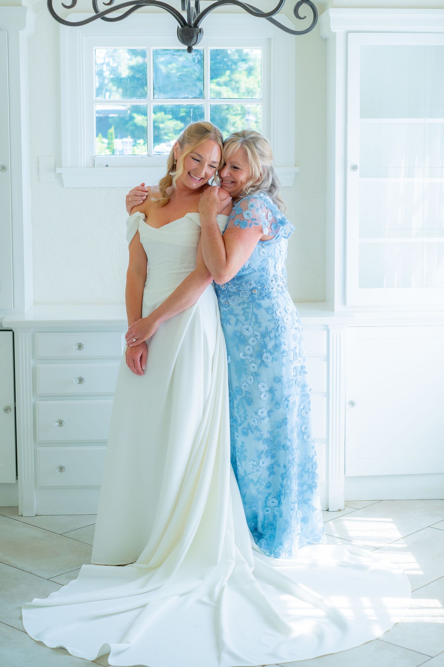 Bride in white off-shoulder gown embraces older woman in blue lace dress in bright white bridal suite.