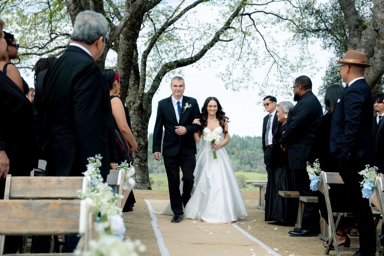 Bride in white gown walks down outdoor aisle with escort past seated guests at a garden wedding ceremony.