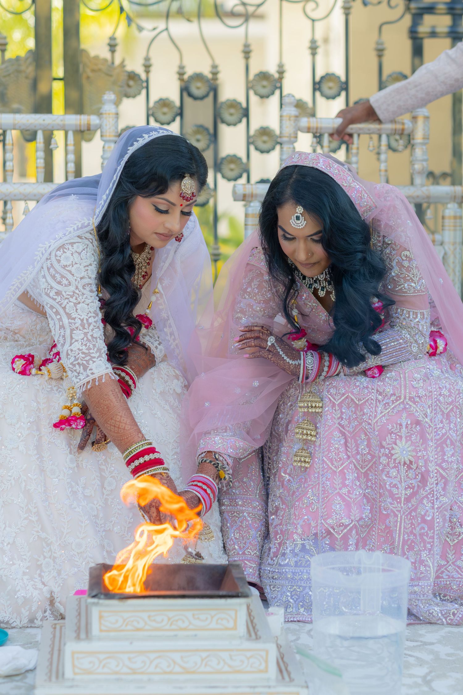 Two brides in white and pink lehengas performing Hindu fire ritual during wedding ceremony.