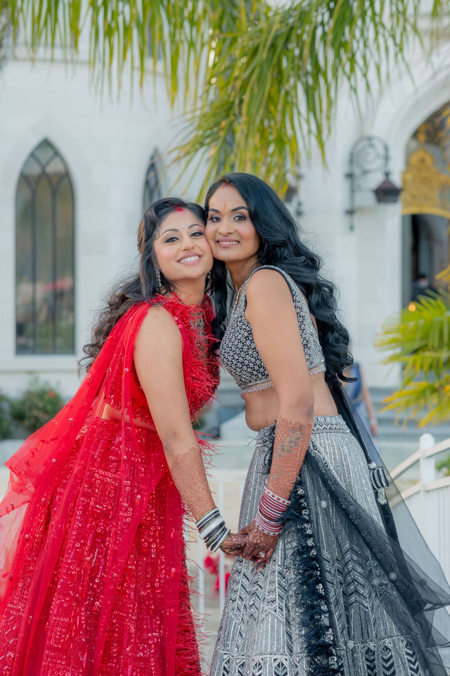Two smiling women in red and grey embroidered Indian lehengas pose together outside a white building.