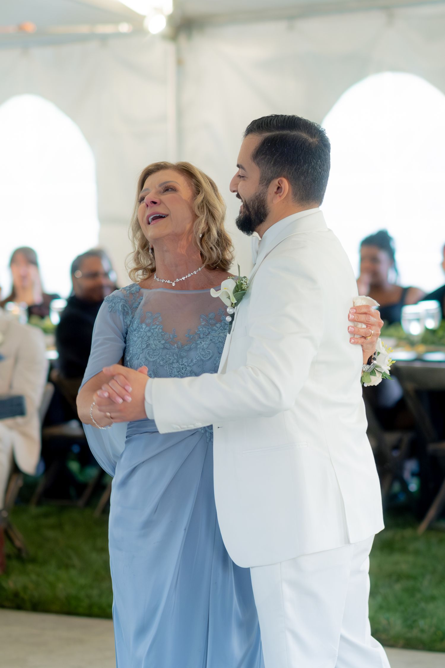 Groom in white suit shares mother-son dance under wedding reception tent with guests.