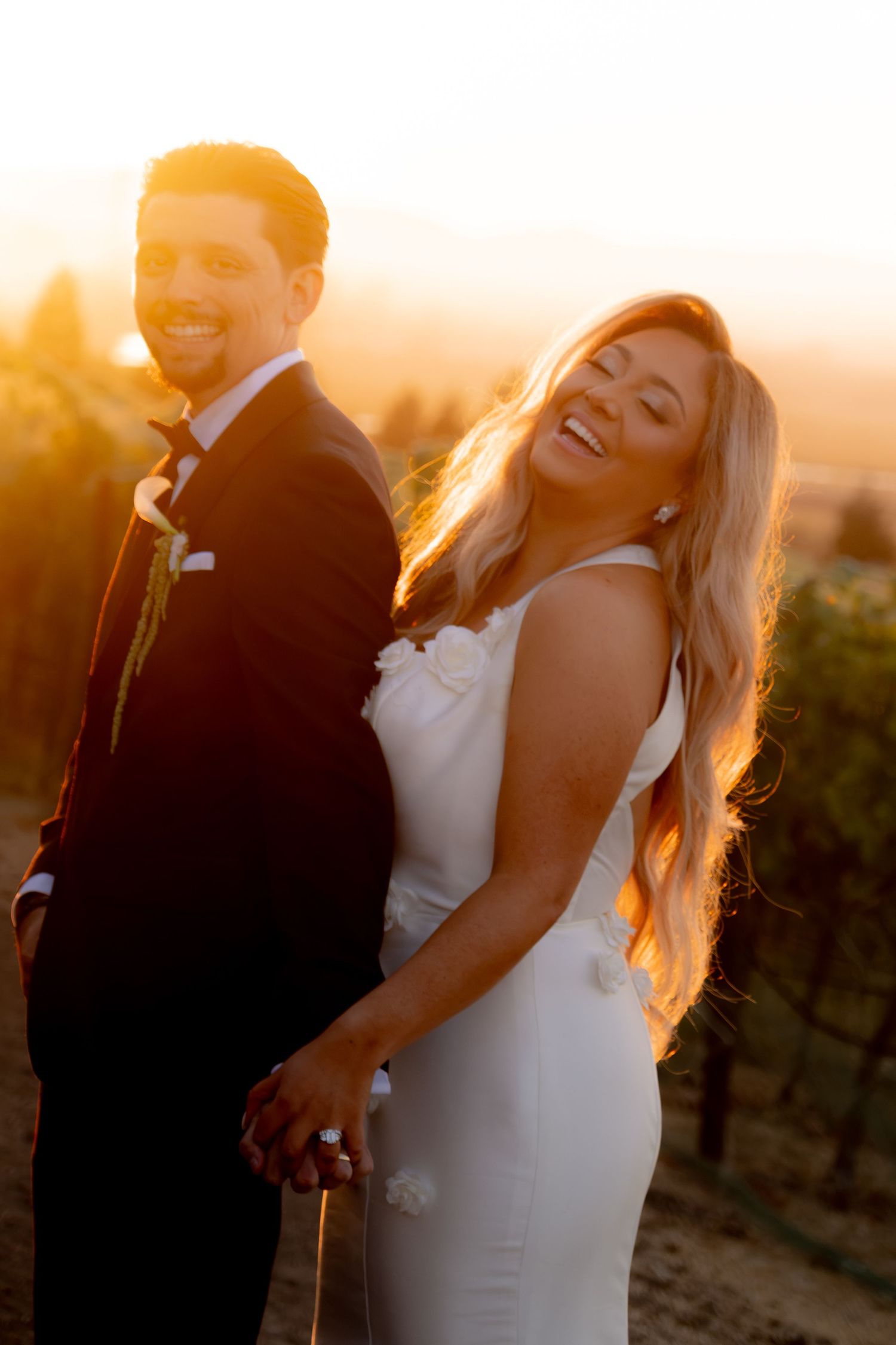 Laughing bride and smiling groom hold hands in a vineyard bathed in warm golden sunset light.