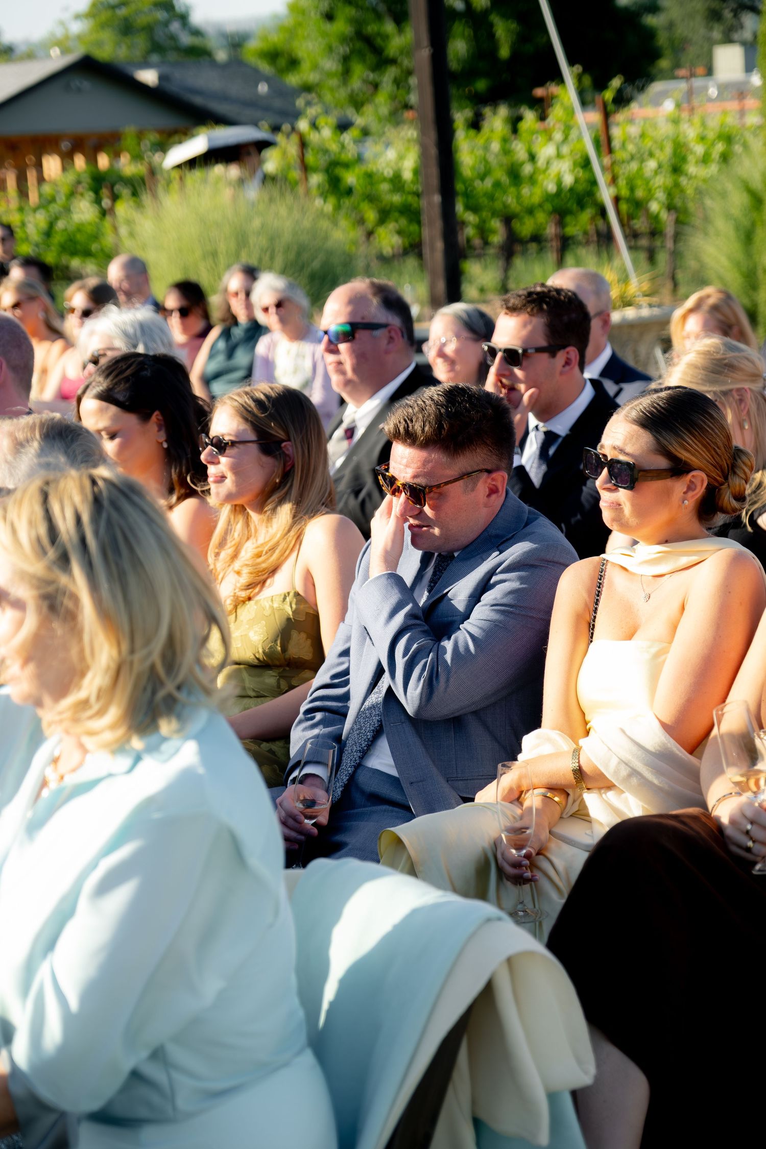 Wedding guests seated outdoors in sunglasses watching an emotional ceremony on a sunny day.