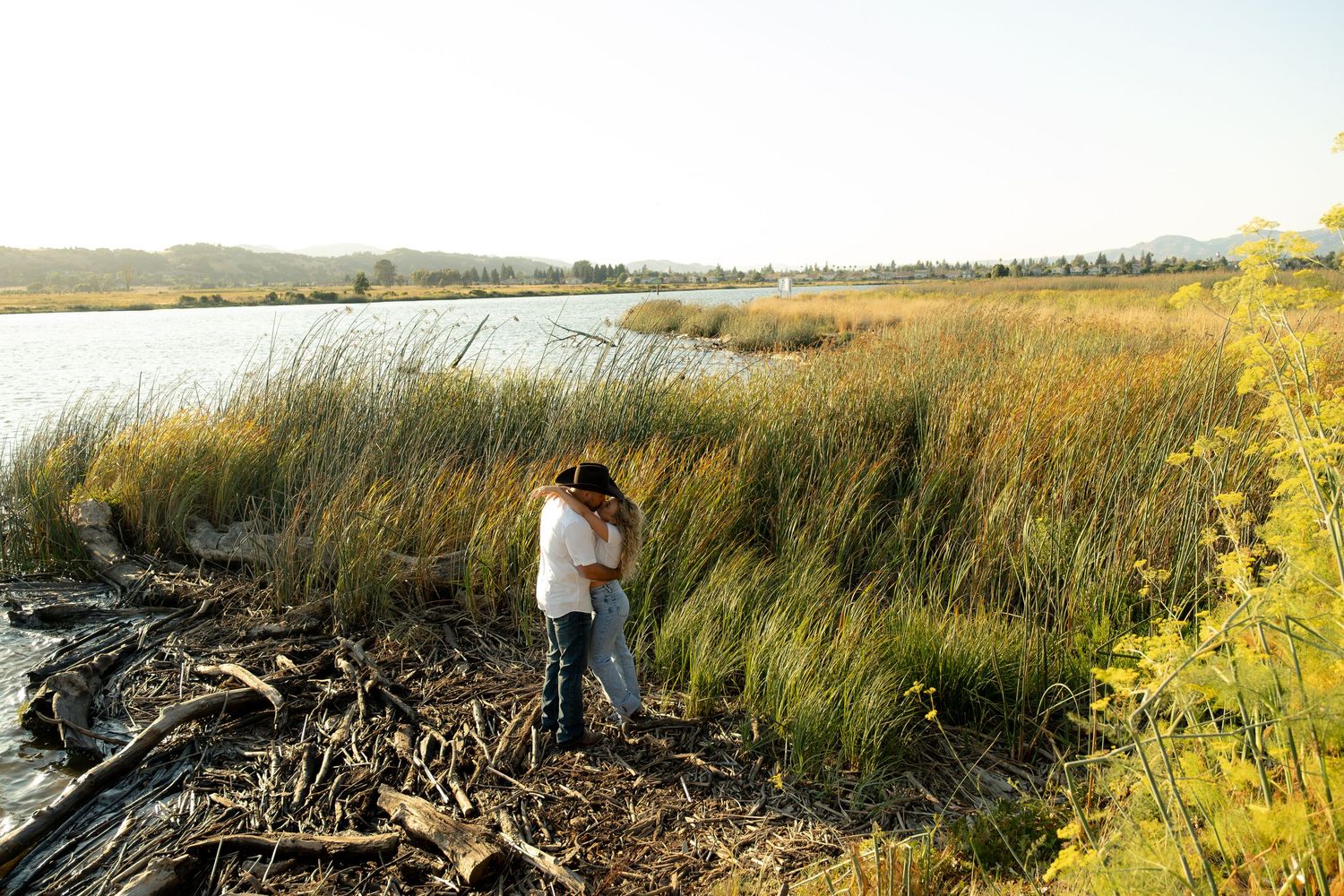 Couple sharing a romantic embrace among tall golden reeds beside a calm lake at sunset.