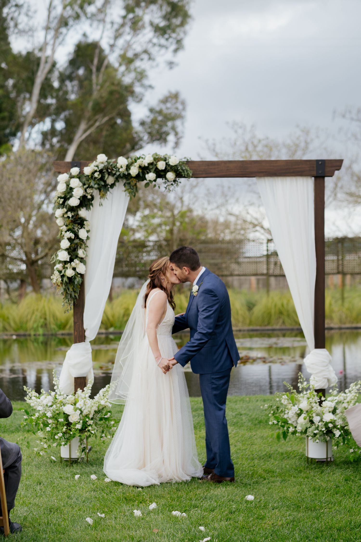 Bride and groom share their first kiss under a wooden arch with white roses beside a serene pond at outdoor ceremony.