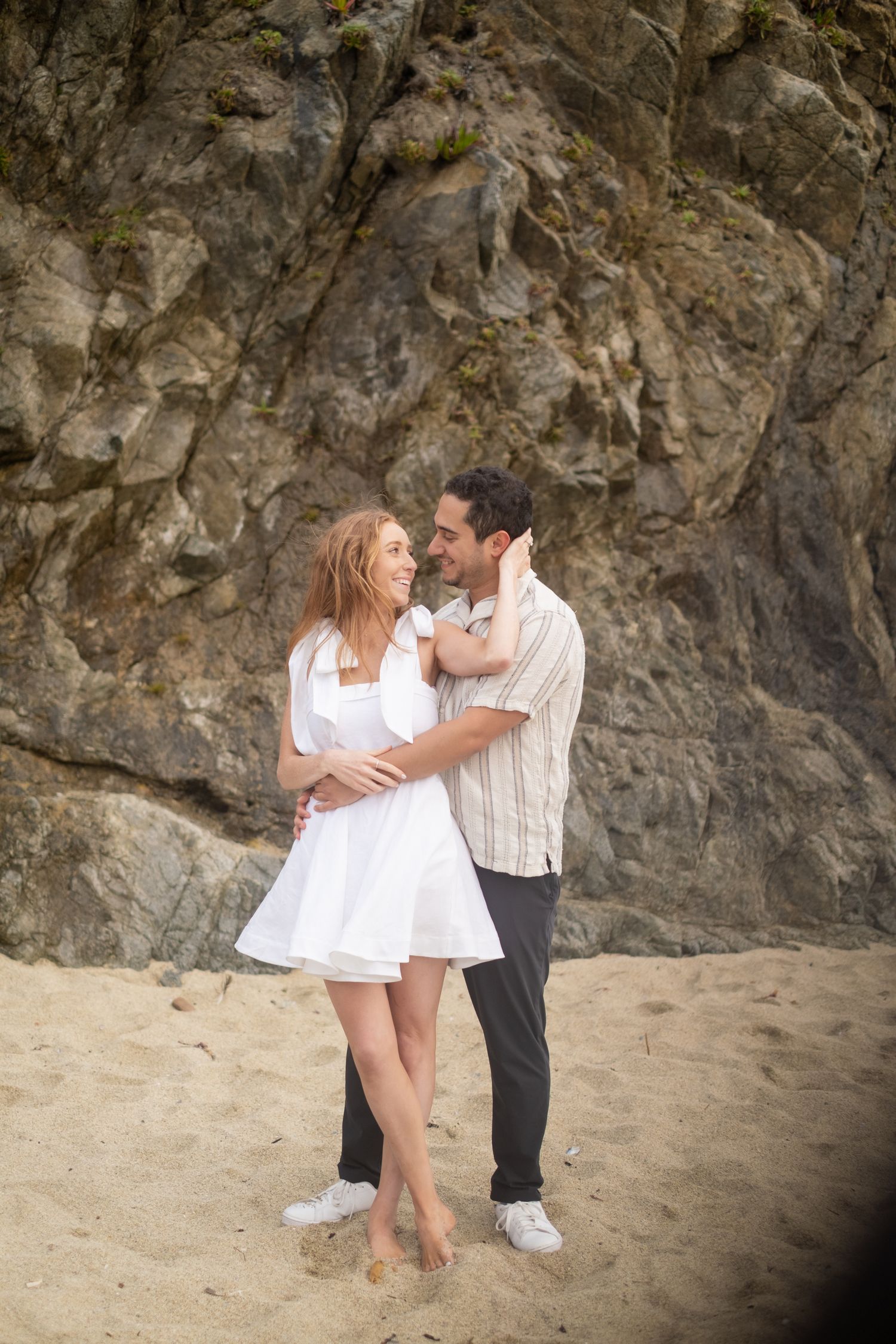 Couple embracing and smiling at each other on sandy beach with large rock face behind.